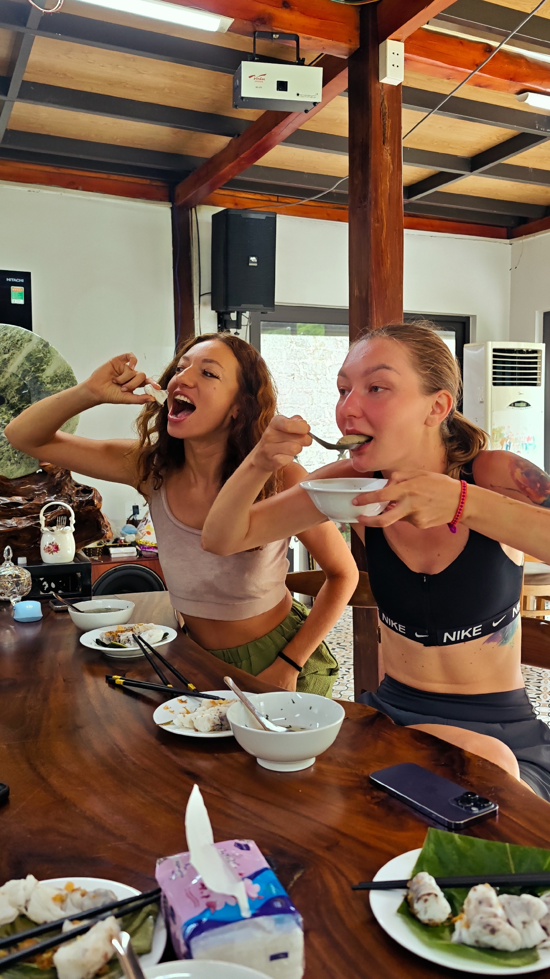 Two women are seated at a wooden table in a room with a partially visible air conditioner. The woman on the left, with curly brown hair, wears a light brown tank top and green shorts. She is in the process of putting food into her open mouth. The woman on the right, with blonde hair pulled back, wears a black Nike sports bra and gray shorts. She is eating from a white bowl with a spoon. The table is laden with white plates holding food items wrapped in green leaves, chopsticks, and white bowls. A smartphone lies near the woman on the right. A tissue box with a white tissue sticking out sits in the foreground. The room has a wooden ceiling and beams with electrical fixtures and a speaker visible. The scene is in Hà Giang 1 Ward, Vietnam.