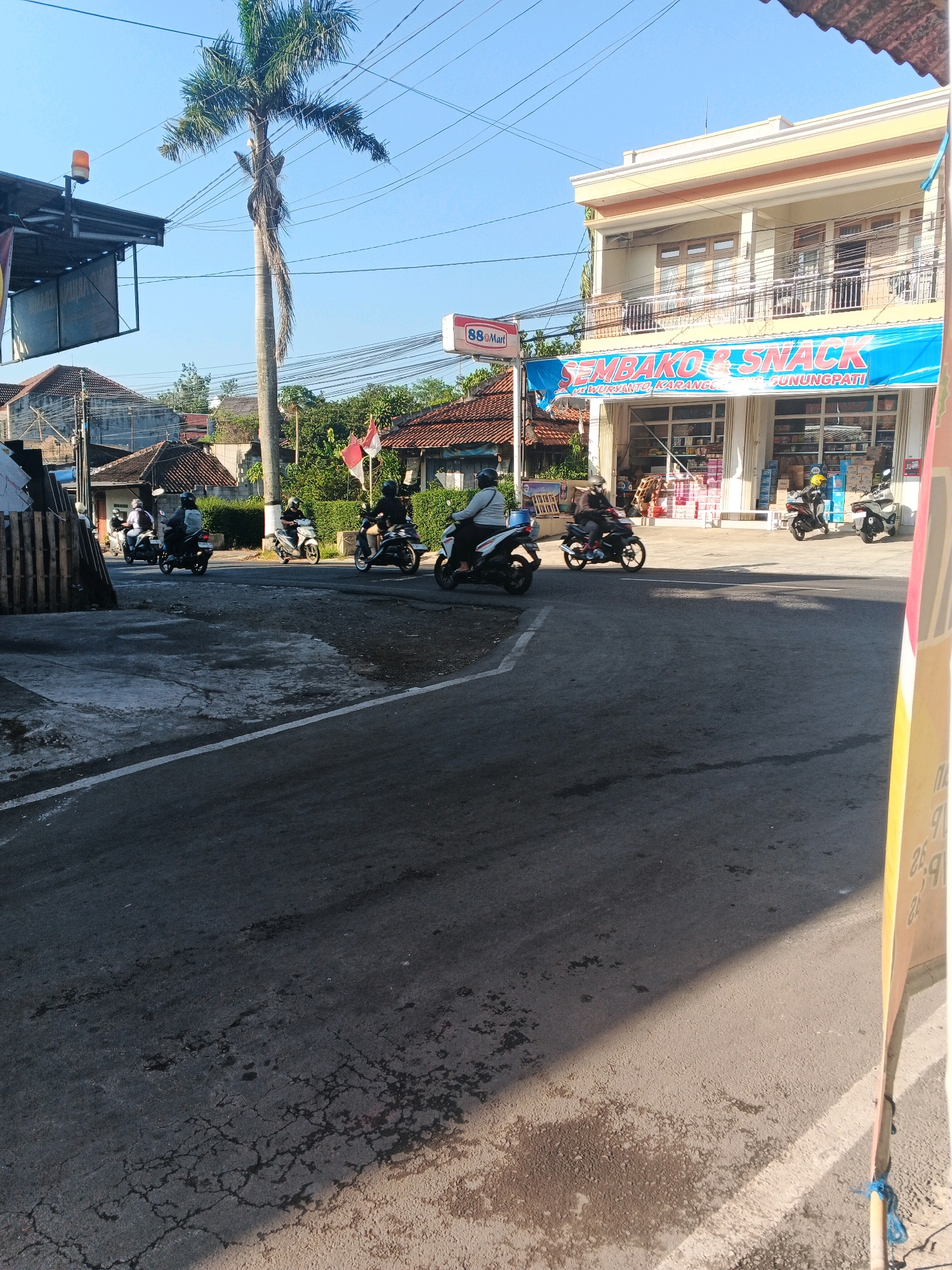 The image depicts a street scene in Semarang, Indonesia, under a clear blue sky. A prominent feature is a tall palm tree with dry fronds. Several motorcycles are in motion on the paved road, with riders wearing helmets, suggesting they are navigating the urban environment. On the right side of the road, a two-story building with a yellow facade houses a "SEMBAKO & SNACK" store. The store's windows display various goods, and several motorcycles are parked outside. Other elements in the scene include power lines, smaller residential buildings with tiled roofs, and some greenery. The road surface shows signs of wear and tear, with some wet patches and cracks.