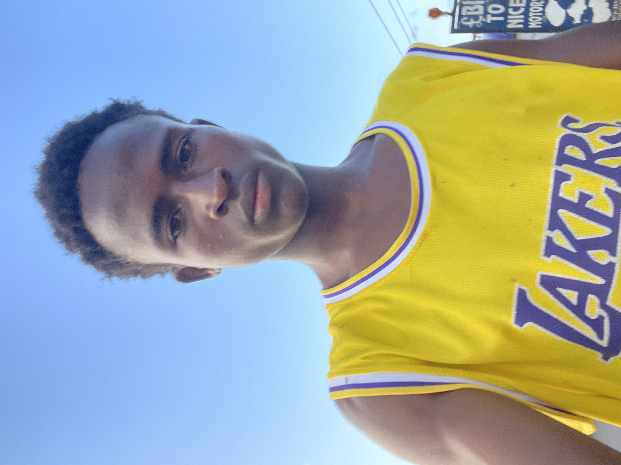 A young Black male is pictured from the chest up, looking directly at the camera. He has short, dark, curly hair and a medium-dark skin tone. He is wearing a bright yellow basketball jersey with purple and white trim around the neckline and armholes. The word "LAKERS" is printed in large purple letters across the front of the jersey. The background is a clear, bright blue sky. To the right of the subject, a portion of a blue and white sign with some text is visible, partially obscured by the subject's shoulder. The sign appears to have some English text that is difficult to read entirely, but possibly includes "TO NICE MOTORS". The overall lighting suggests it is daytime, likely outdoors. The environment appears to be an urban or semi-urban setting based on the sign, possibly on a street or roadside. Specific details about Jos, Nigeria are not discernable from the image itself.