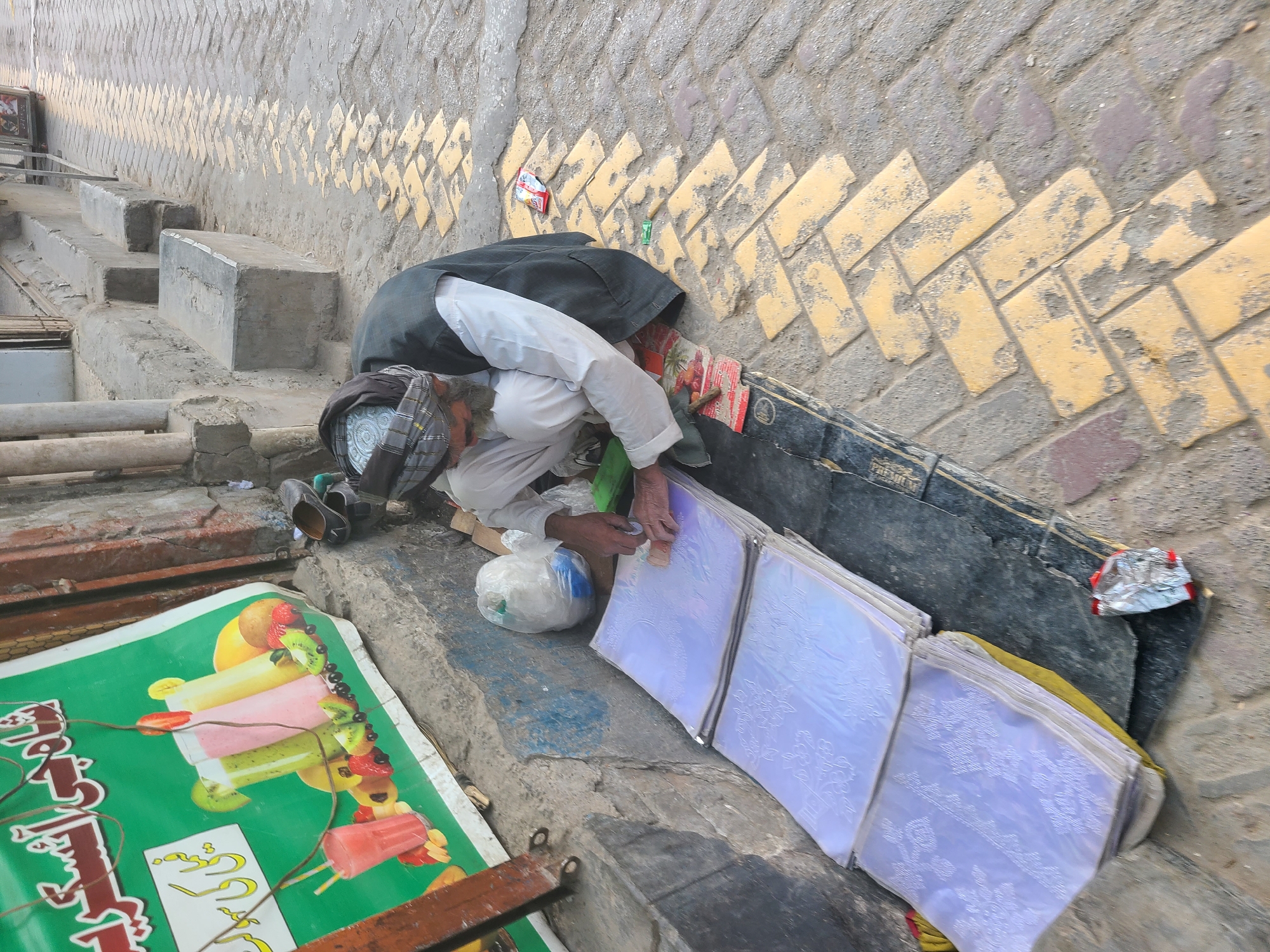 The image shows a man bending over beside a wall in Quetta City Tehsil, Pakistan. The wall is made of tan bricks arranged in a herringbone pattern, with a gray concrete top. The man is wearing a dark jacket, white shirt, and a patterned headdress. He appears to be working or arranging items on a ledge next to the wall. These items include a plastic bag and a stack of what appears to be white, patterned papers or cloths. To the left of the man is a colorful sign with images of ice cream and juice. There are concrete steps leading up next to the ledge where the man is working. Debris and litter are scattered around the area.