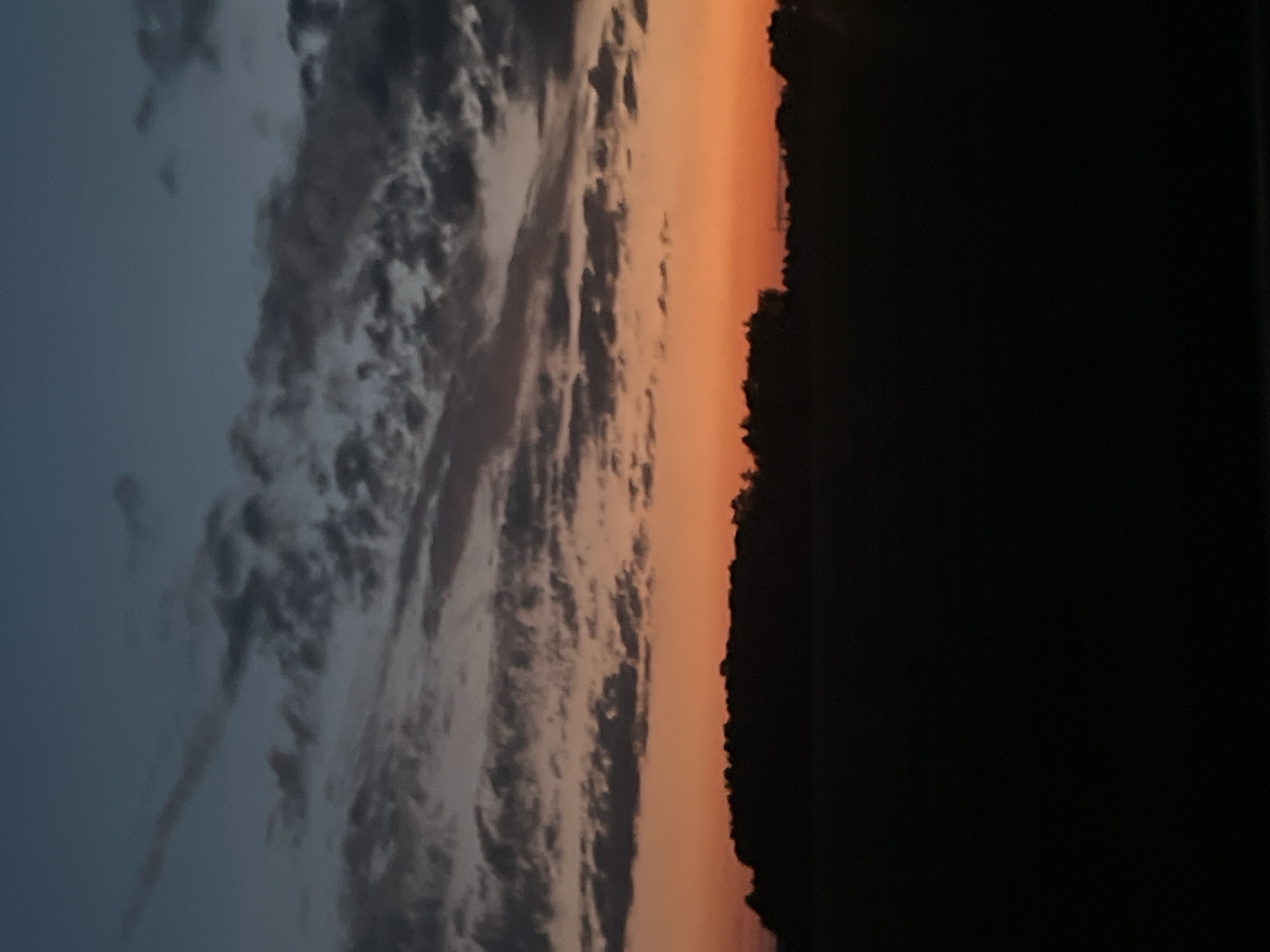 The image depicts a silhouetted landscape at dusk. A dark, treed horizon line cuts across the lower third of the frame. Above this, a vibrant band of orange transitions into a deep blue sky filled with scattered, wispy clouds. The scene is devoid of any human presence or identifiable structures, suggesting a rural or natural setting. The location is specified as Поварёнки, Russia.