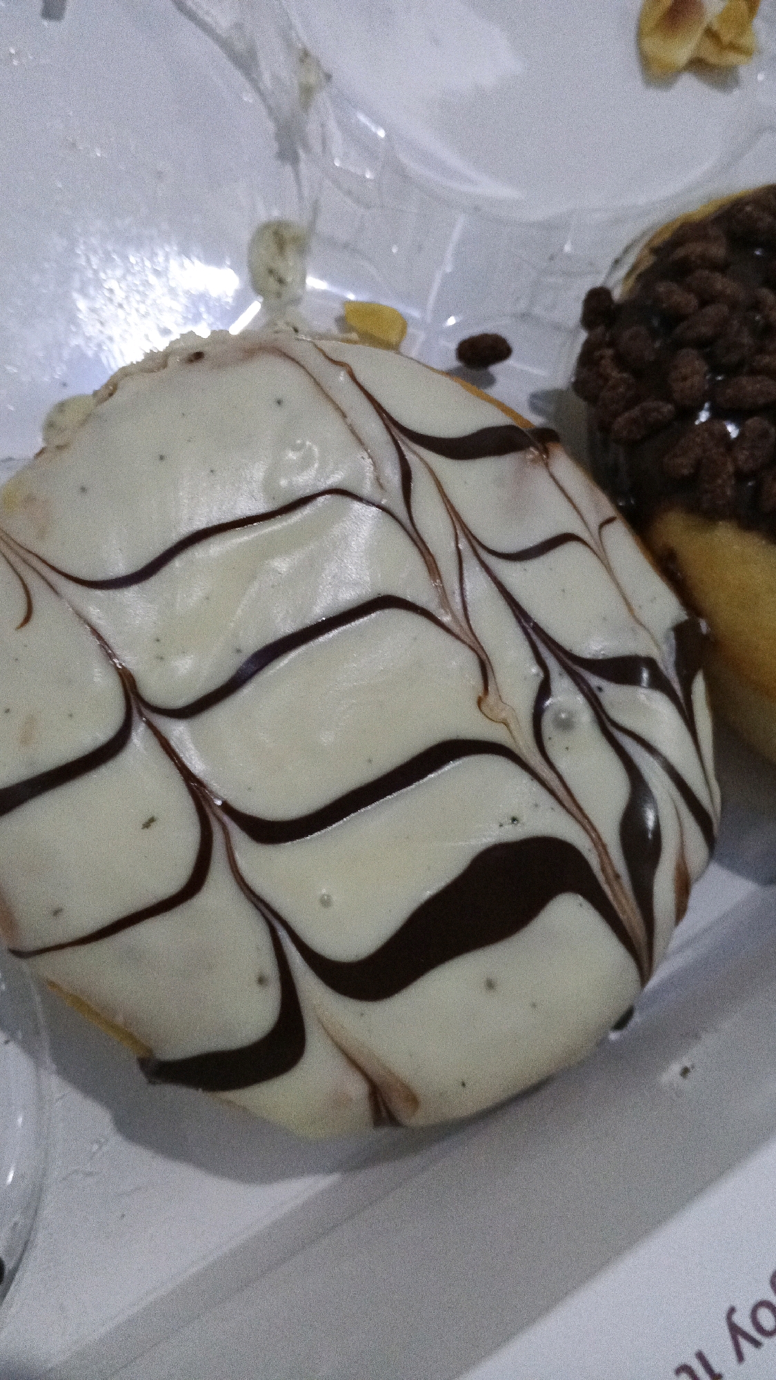 The image shows two donuts. One donut is in the foreground, covered in white icing with dark brown chocolate drizzled in a marbled pattern. The second donut, partially visible in the background, is covered in chocolate icing and topped with small, crunchy, chocolate-flavored pieces, possibly cereal or nuts. The donuts are in a white paper container, with some plastic wrapping visible. The scene appears to be indoors, with artificial lighting. The image does not contain any explicit information indicating the location in Semarang, Indonesia.