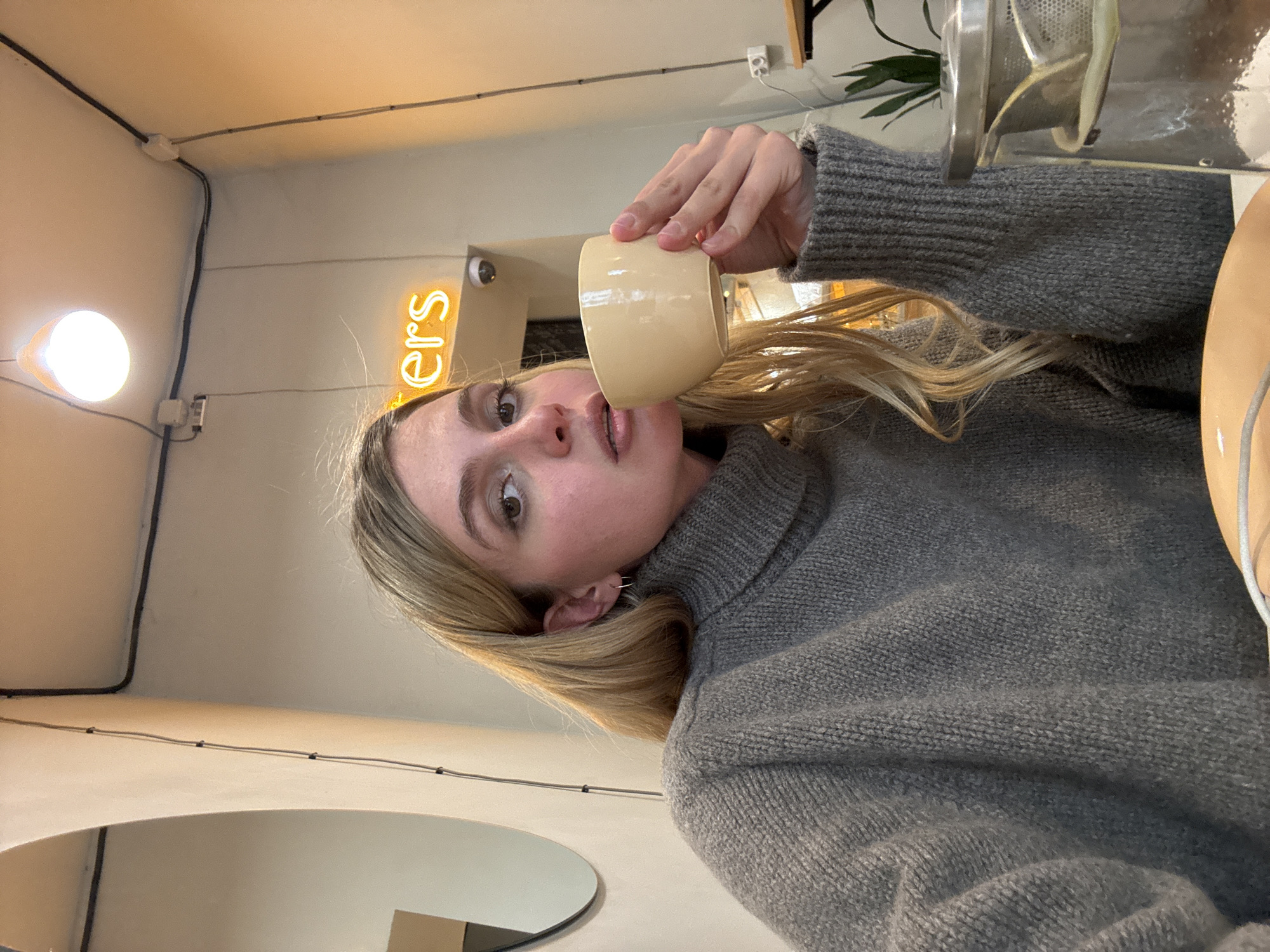 A young, fair-skinned woman with blonde hair is holding a light-colored mug and drinking from it. She is wearing a grey turtleneck sweater. The background includes a white wall, a neon sign reading "ers", a light fixture, and a partial view of a round mirror. The scene appears to be indoors, possibly a cafe or restaurant. Without additional context or clues, it is impossible to verify that the location is Tbilisi, Georgia.
