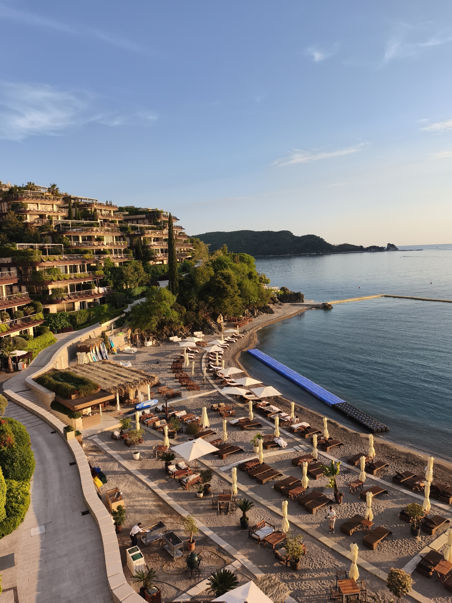 This high-angle shot captures a scenic coastal resort in Budva, Montenegro, bathed in the warm, golden light of either early morning or late afternoon. The scene features a terraced hillside adorned with multi-story, modern buildings, likely hotel rooms or luxury apartments, integrated seamlessly with lush green landscaping, including numerous trees and shrubs. Each level of the buildings boasts wide balconies, many also greened with plants.
Below the hillside, a sandy beach stretches along the calm, blue-green sea. The beach is meticulously arranged with numerous wooden sun loungers, many accompanied by white parasols. Small tables are placed between seating arrangements. A thatched-roof structure, likely a beach bar or restaurant, stands prominently on the sand, surrounded by more seating areas. Nearby, several paddleboards or surfboards are stacked, suggesting water sports activities.
A wide, paved walkway curves along the base of the hillside, separating the resort buildings from the beachfront. In the beach area, at least two individuals are visible; one person, possibly staff, is pushing a cart on the sand.
The sea itself is calm, with a long, bright blue floating dock or walkway extending into the water parallel to the shore. A natural stone pier also juts out into the water further down the coast. Distant hills and landmasses line the horizon across the bay. The sky is a clear, bright blue with a few wispy clouds, indicating pleasant weather. On a blue board near the beach bar, the text "ADIMAR" is visible.