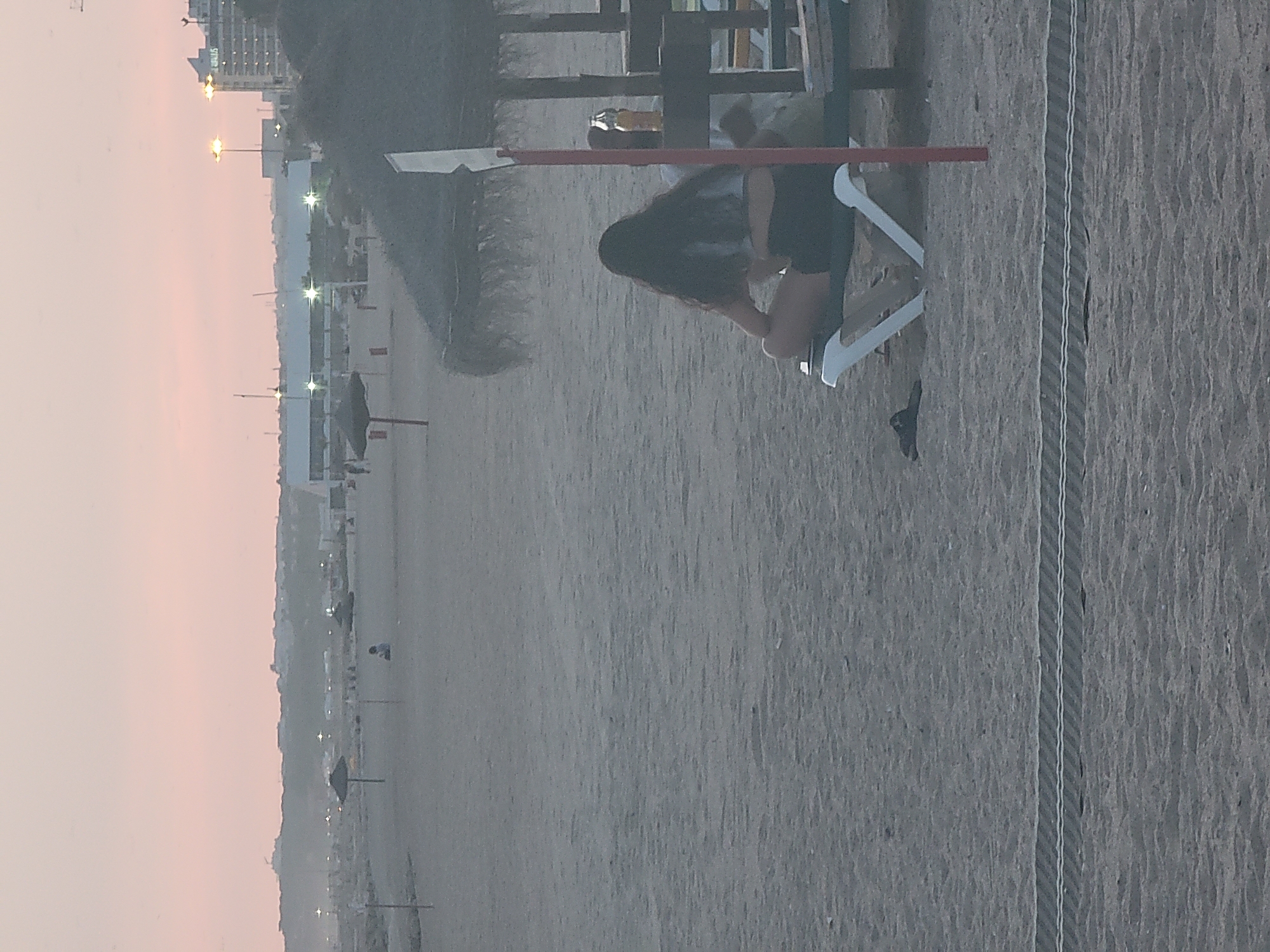 This image depicts a beach scene in Quarteira, Portugal, during what appears to be dusk or early morning, indicated by the soft, pinkish hue of the sky. In the foreground, a person with long dark hair sits on a white lounge chair facing away from the camera. They are wearing a light-colored top and dark shorts. Next to the lounge chair, a pair of dark flip-flops rests on the sand. In the background, several thatched-roof umbrellas are spread across the sandy beach. Beyond the umbrellas, buildings and streetlights are visible along the coastline, suggesting a developed urban area. The overall atmosphere is tranquil and relaxed.