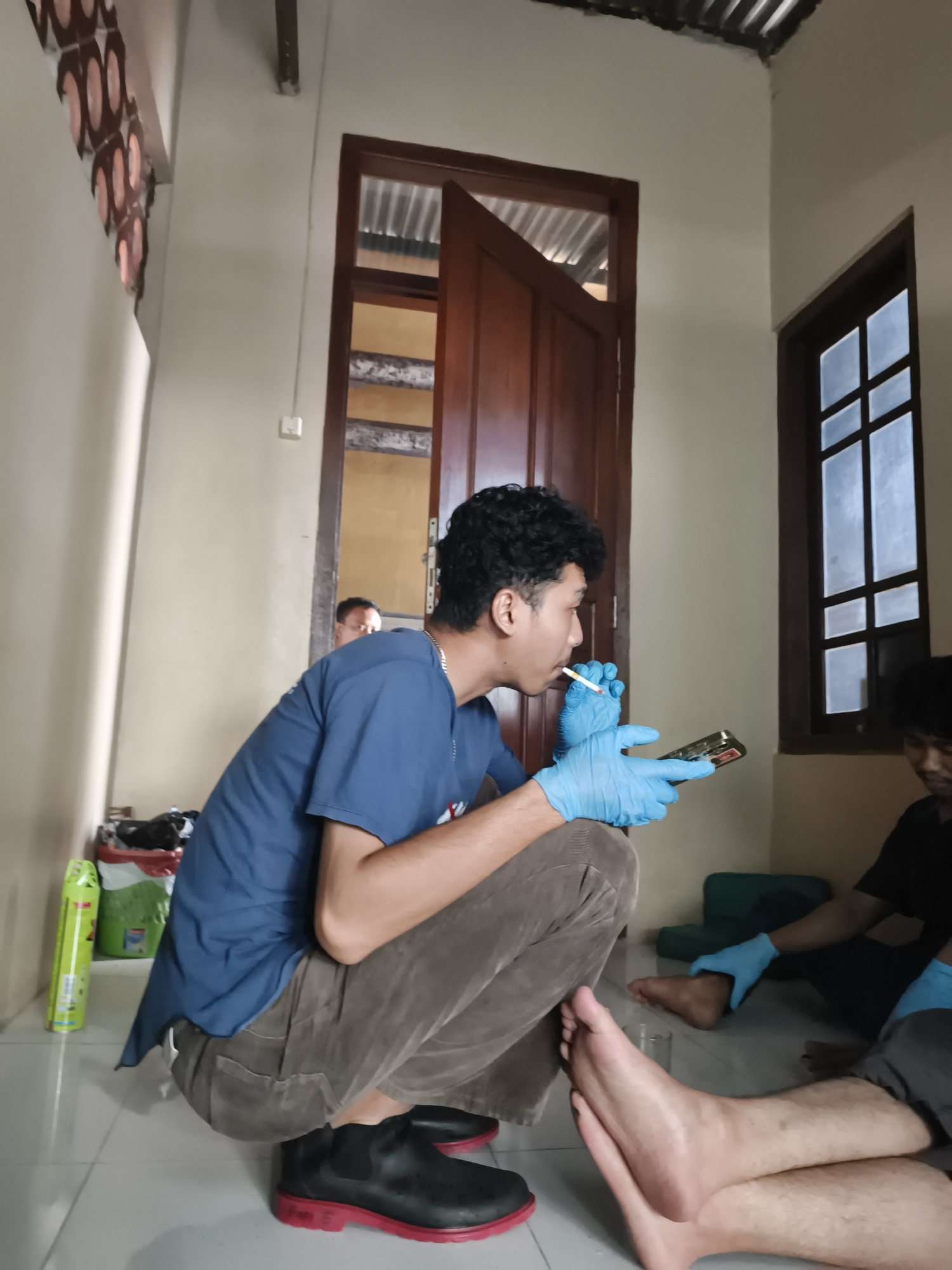 The image depicts an indoor scene, likely a dwelling, in Pekalongan, Indonesia. A young man wearing a blue t-shirt and brown pants is crouching, holding a cigarette in his mouth and a mobile phone in his gloved hands. Another person, partially visible, is seated on the floor, also wearing gloves, and appears to be attending to the first man's foot. The room has tiled flooring and a wooden door and window are visible. Decorative elements are present on the wall, and various objects, including a spray can and bags, are scattered around. The overall atmosphere suggests a casual or possibly medical-related gathering.
