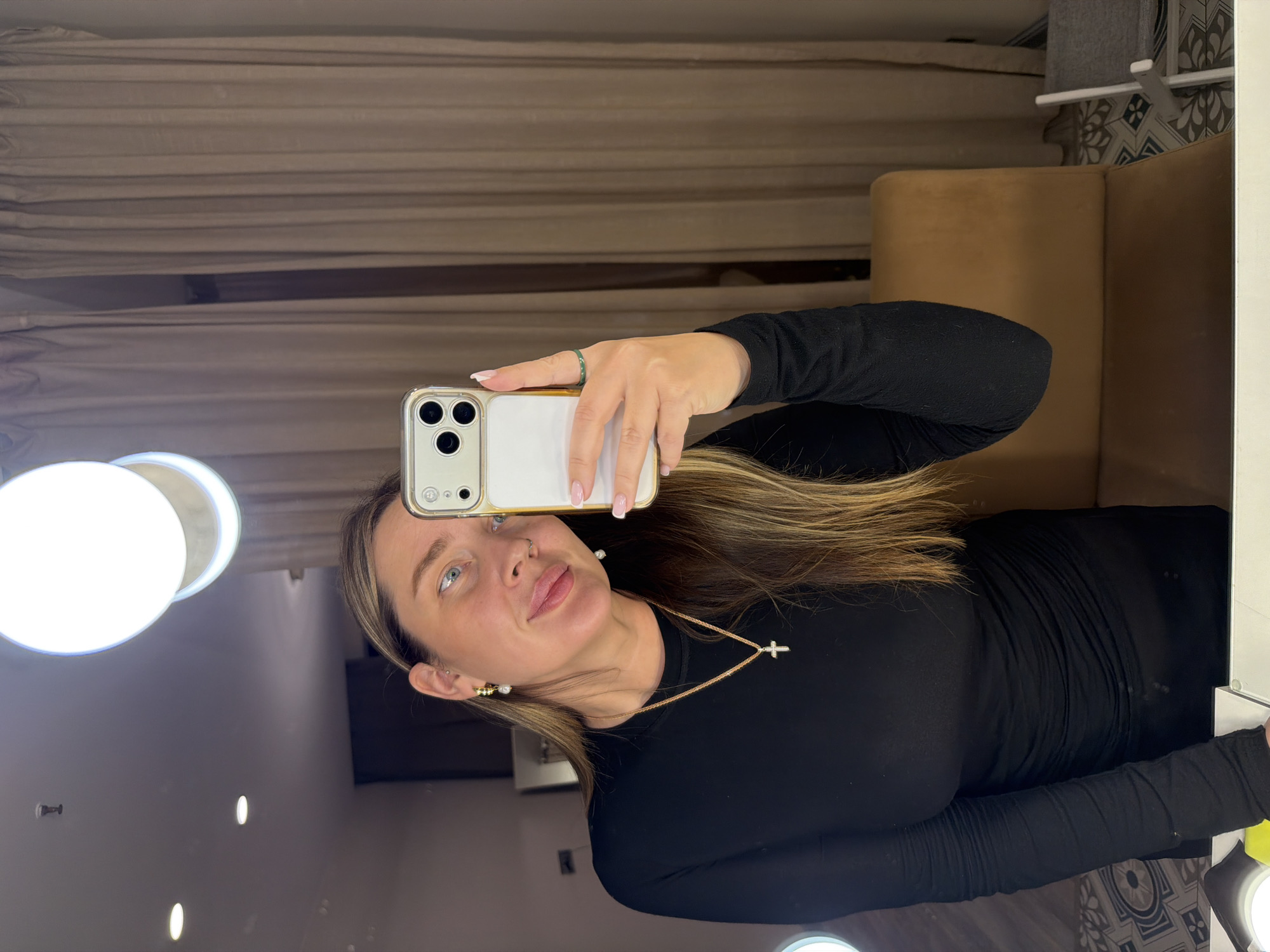 A woman is taking a selfie in a mirror. She has blonde hair, blue eyes, and is wearing a black long-sleeved shirt and a gold necklace with a cross pendant. She is holding a white and gold iPhone. The mirror is surrounded by lights, and there is a beige curtain and chair in the background. The scene does not contain any details that indicate it is specifically located in Moscow, Russia.