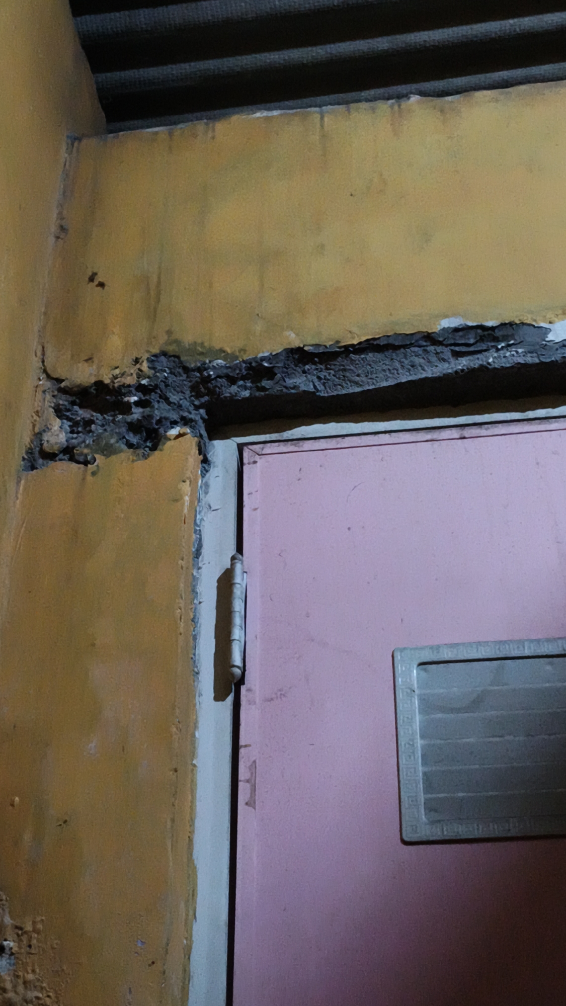 The image depicts a close-up view of a damaged doorframe and a pink door in Kerep Kidul, Indonesia. The doorframe shows significant structural damage, with crumbling plaster and exposed concrete along the top and left sides. The plaster is discolored with dark streaks and stains. Above the doorframe, a section of a dark, ribbed ceiling is visible. The pink door is slightly ajar, revealing a hinge on the left side. A light-colored rectangular panel with a textured border is affixed to the right side of the door. The overall impression is one of disrepair and neglect.