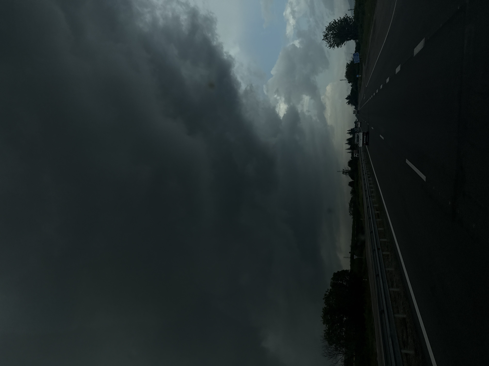 The image depicts a highway adjacent to what appears to be a rural area in Zlatina, Bulgaria. The highway stretches into the distance, with a metal guardrail visible on the side. Several cars are visible on the road. The sky is dominated by dark, ominous storm clouds, with a small patch of lighter clouds visible.