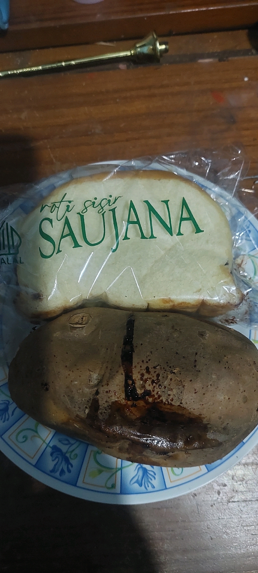 The image depicts two food items on a patterned plate, set on a wooden surface. The upper item is a loaf of bread, possibly a sliced loaf, encased in clear plastic packaging. The bread has a light brown crust and a soft interior. Green text printed on the plastic packaging reads "roti sisir SAUJANA". To the left of the text, a symbol resembling a building or a structure is visible, followed by the word "HALAL". The lower item is a dark brown, oval-shaped food item, possibly a baked potato or a type of bread, with a cracked surface revealing a dark, glistening interior. The plate has a white base with a repeating pattern of blue floral designs and square borders. In the background, a golden object with a decorative top, possibly a piece of brass hardware or an ornate pen, rests on the wooden surface. The lighting suggests an indoor setting. The scene is presented in a top-down perspective. There are no people visible in the image. The image was taken in Lumajang, Indonesia.