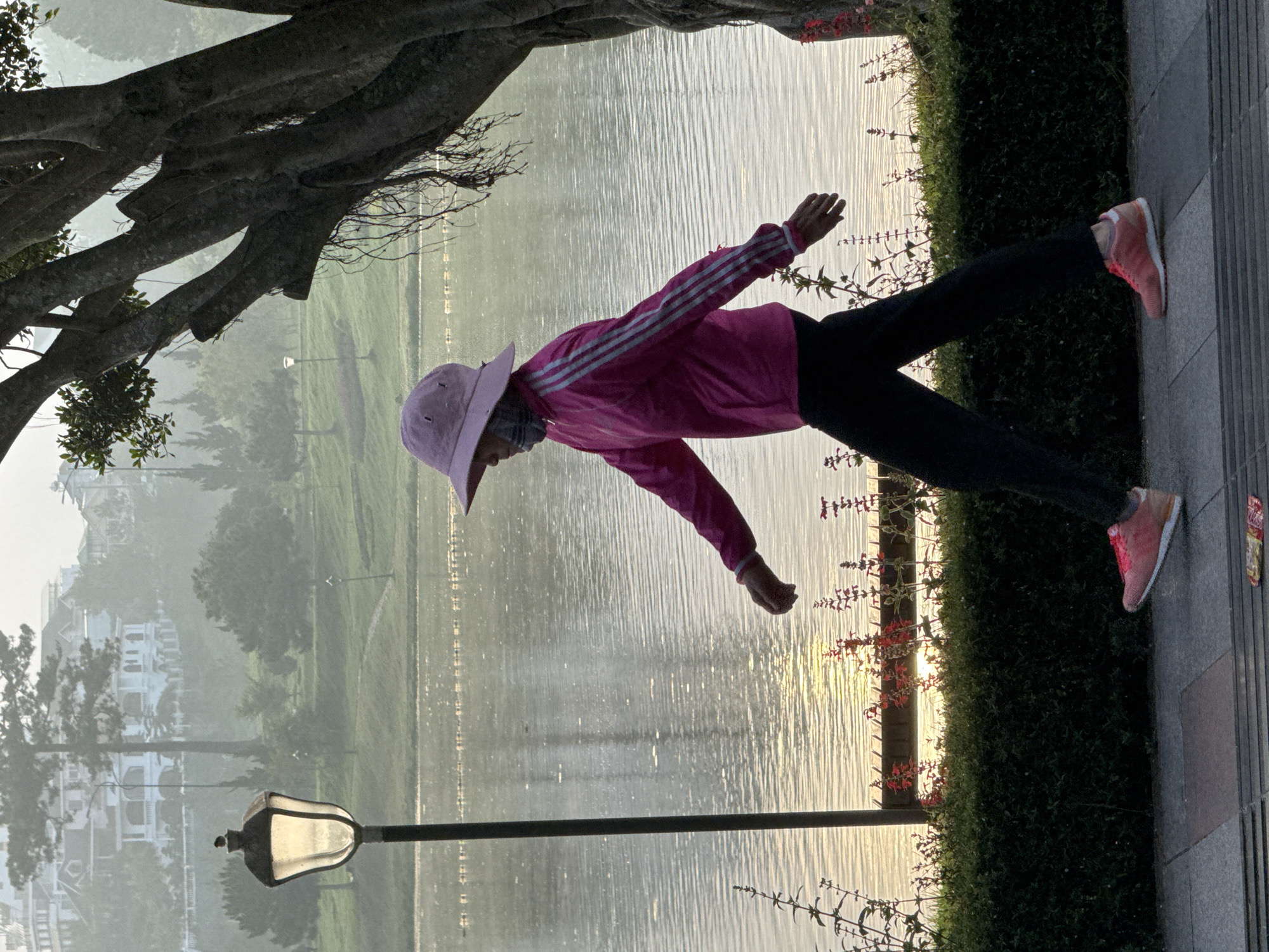 An individual walks along the paved pathway beside Xuân Hương Lake in Đà Lạt, Vietnam. The person is wearing a pink wide-brimmed hat, a pink hooded jacket with white stripes on the sleeves, black pants, and pink athletic shoes. Their face is partially obscured by a patterned face covering. The individual is in profile, facing left, with their arms in a natural walking posture.
To the left of the individual is a lamppost with an illuminated lantern. In the background, across the lake, is a hazy landscape featuring rolling green hills, trees, and buildings, indicative of the city's mountainous terrain. The lake surface is calm, reflecting the soft, diffused light of what appears to be early morning or late afternoon. A line of manicured green bushes and flowering plants with red blossoms borders the pathway and the lake. A large tree with thick branches and hanging roots is positioned to the right of the frame, partially obscuring the background. The pathway is made of large, rectangular paving stones. A piece of discarded wrapper lies on the ground in the lower right corner of the image.