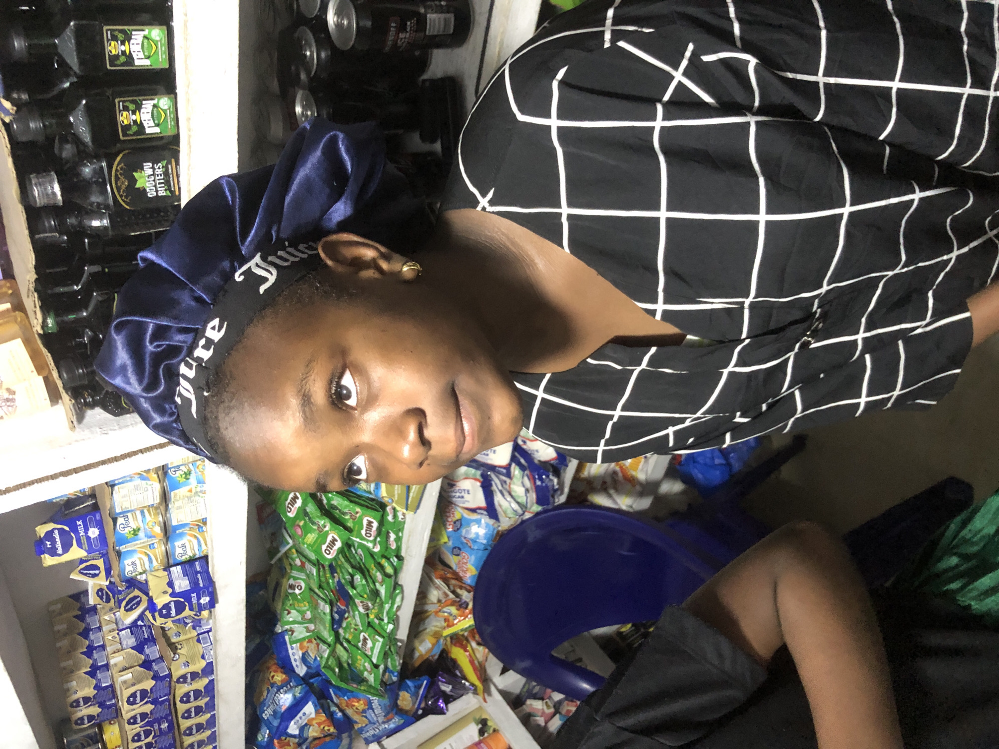 The image depicts a young Black female wearing a blue satin bonnet with "Juicy" written in white on the band. She has gold hoop earrings and is looking upwards and to her right. She is wearing a black and white checkered shirt. The background shows shelves stocked with various products, including milk cartons, small bottles of what appears to be alcohol, and packets of snacks. To the left of the female, another person's arm and shoulder are visible, wearing a black t-shirt. A blue plastic chair is positioned behind the female. The setting appears to be a small shop or convenience store in Ado, Nigeria, given the presence of branded products commonly found in such establishments. The lighting suggests an indoor environment, possibly with artificial illumination.