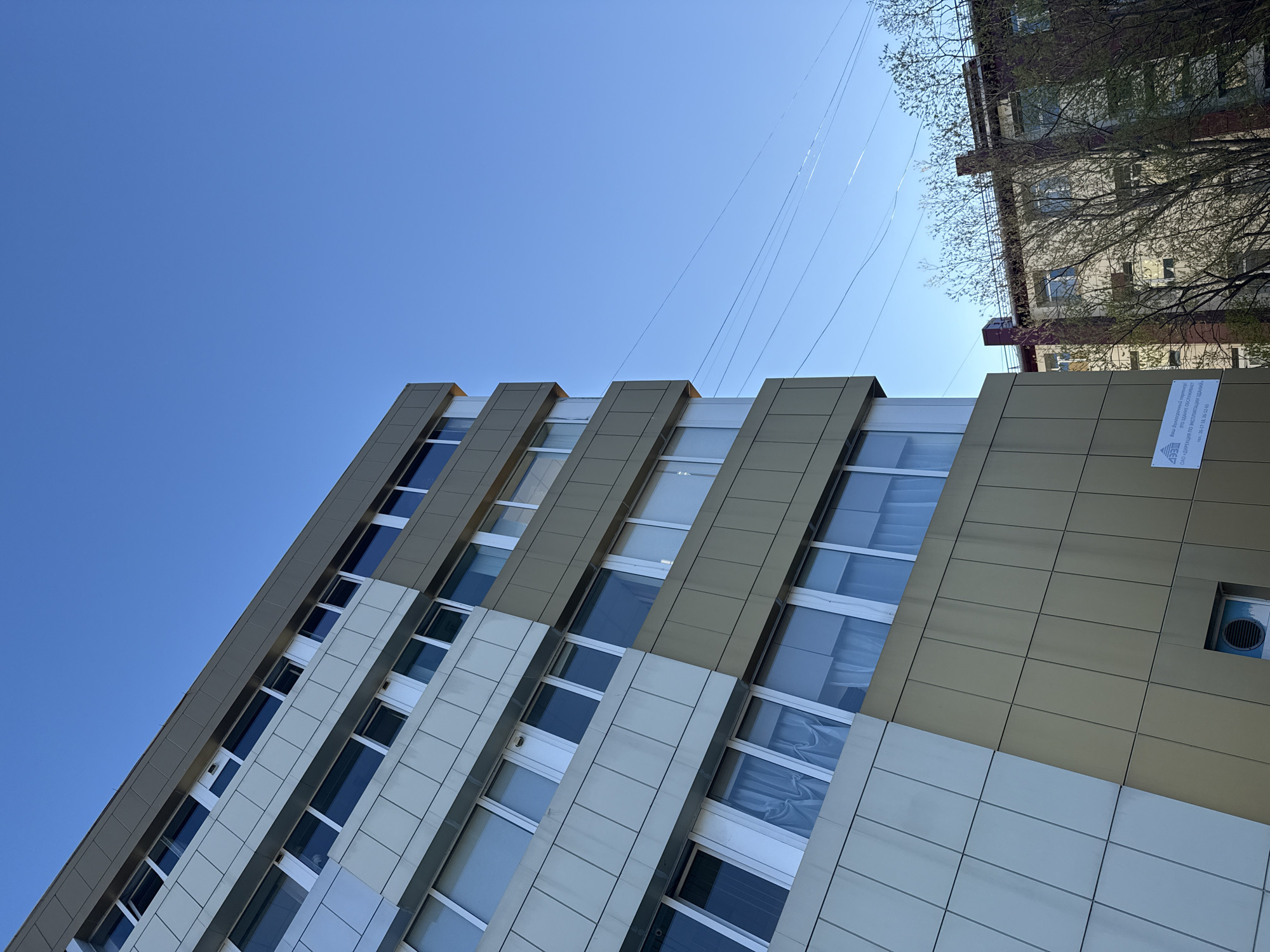 A low-angle, outdoor daytime shot captures a modern, multi-story building against a clear blue sky with visible contrails. The building has a distinct architectural design, featuring alternating panels of light beige and gold-toned cladding, with vertical strips of windows.
On the right side of the frame, a portion of an older building is visible, showing a facade with dark accents and emerging tree branches with spring foliage.
Attached to the right side of the modern building, there is a sign with text in Russian. The text reads "Ваш долгосрочный партнер! Это здание обслуживает ОАО «ДИРЕКЦИЯ ПО ЭКСПЛУАТАЦИИ ЗДАНИЙ» тел: 26-17-18, 26-12-45" indicating that the building is serviced by "JSC 'Building Management Directorate'".
The location is likely Petropavlovsk-Kamchatsky, Russia, based on the Russian language on the sign and general architectural context.