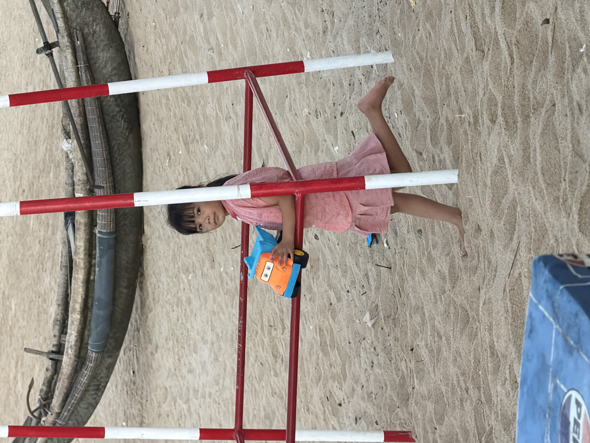A young Asian girl wearing a pink dress is hanging upside down from a red and white striped metal structure on a sandy surface. She is holding a blue and orange toy car. The location appears to be a beach or sandy area in Thuận An Ward, Vietnam, given the presence of sand and a glimpse of a blue object possibly related to the beach environment. There are no other people visible in the frame.