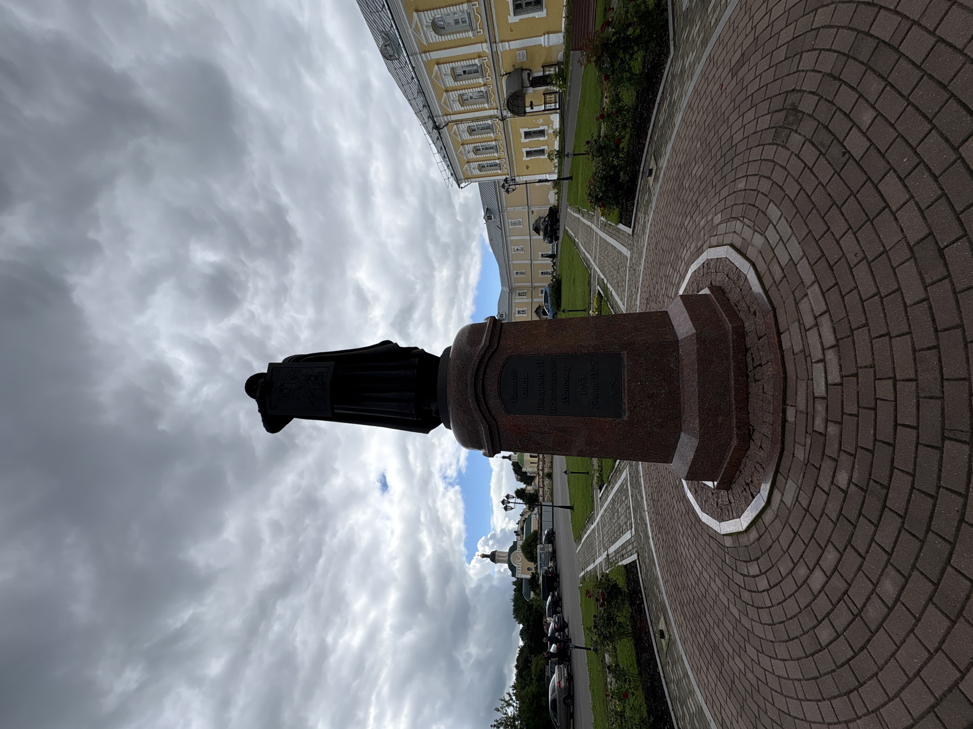 A dark, silhouetted statue stands on a stone pedestal in a public square paved with red bricks. The statue depicts a robed figure holding a book, with its head bowed. The pedestal is inscribed with Cyrillic text and dates. In the background, a yellow building with a gray roof and white trim stretches across the scene. Further in the distance, a church with a green dome and a golden cross is visible. Cars are parked on the side of the road, and a few people are walking. The sky is overcast with dramatic, gray clouds. The location is Sof'ino, Russia.