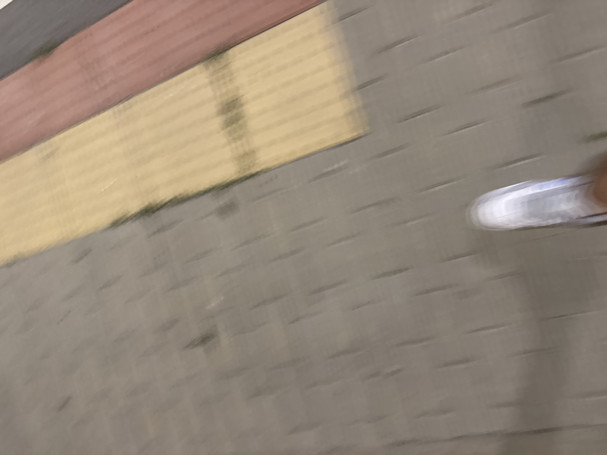 The image shows a blurry view of a person's foot wearing a white athletic shoe walking on a paved surface. The paving stones are a light gray color and laid in a diamond pattern. To the right of the gray paving stones are three parallel strips of different colored paving stones: yellow, red, and dark gray. The scene appears to be outdoors. The location is identified as Shumen, Bulgaria.