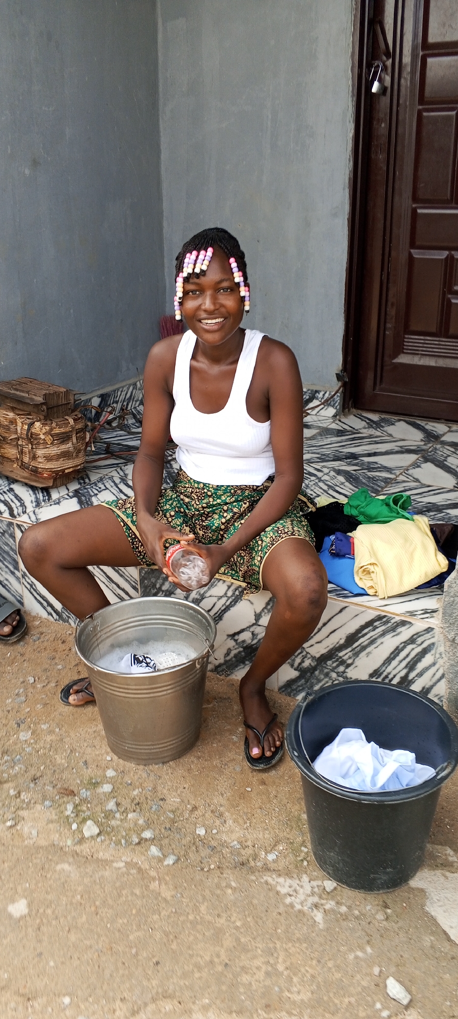 A young woman with braided hair adorned with colorful beads sits on the steps of a building in Unknown, Unknown. She wears a white tank top and a patterned skirt. A silver bucket filled with soapy water sits between her legs, and she holds a plastic water bottle. A black bucket containing white clothing rests on the ground to her right, and a pile of folded clothes is visible behind her. The steps are tiled, and a stack of objects wrapped with rope sits to her left. The ground in front of her is dirt with scattered pebbles.