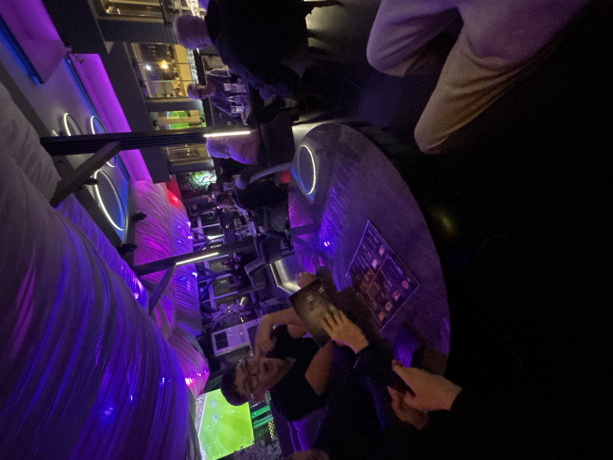 The image depicts an interior scene, likely a bar or lounge in the city of Zlatograd, Bulgaria. Several people are present, some sitting at a round table illuminated with a ring of light. A person is viewing a menu while another person is looking at the camera. There are other people at other tables in the background. A large, illuminated art installation dominates the left side of the scene. A screen is visible in the lower left corner, displaying what appears to be a soccer game. The lighting is dim and primarily purple.