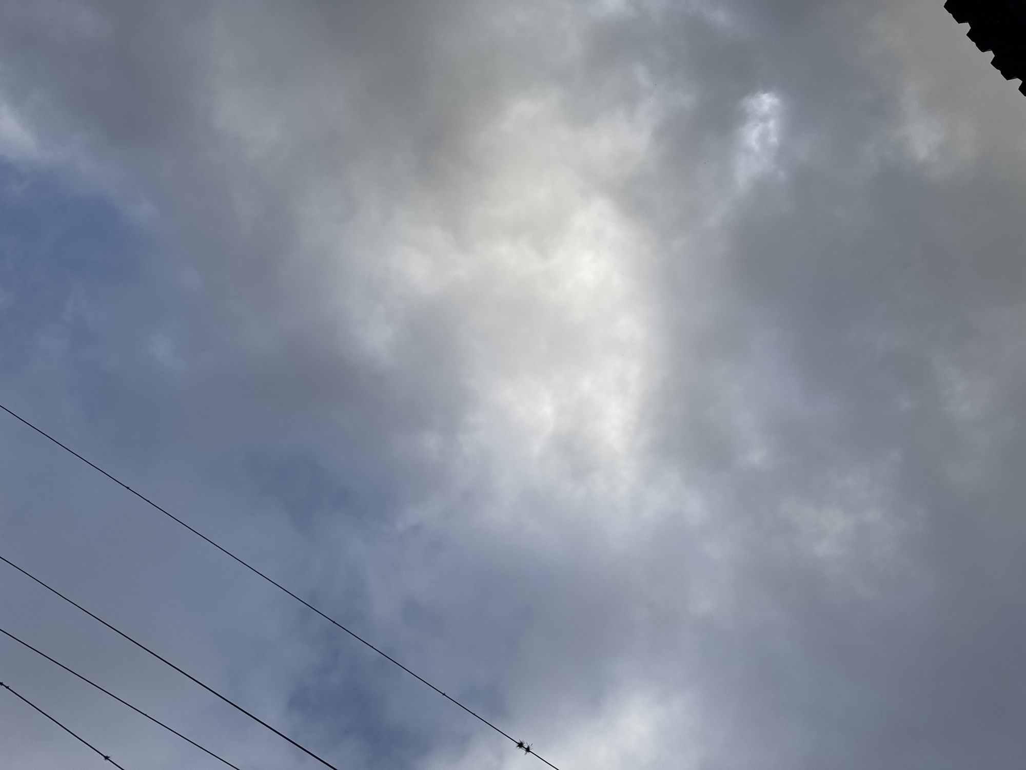 The image depicts a cloudy sky with overcast conditions. The clouds are primarily gray and white, suggesting a potentially stormy or rainy day. A set of power lines diagonally crosses the lower-left portion of the image. There is a hint of a dark structure, possibly a roof, in the upper-right corner. The location is potentially the city of Parroquia Ocumare, Venezuela.