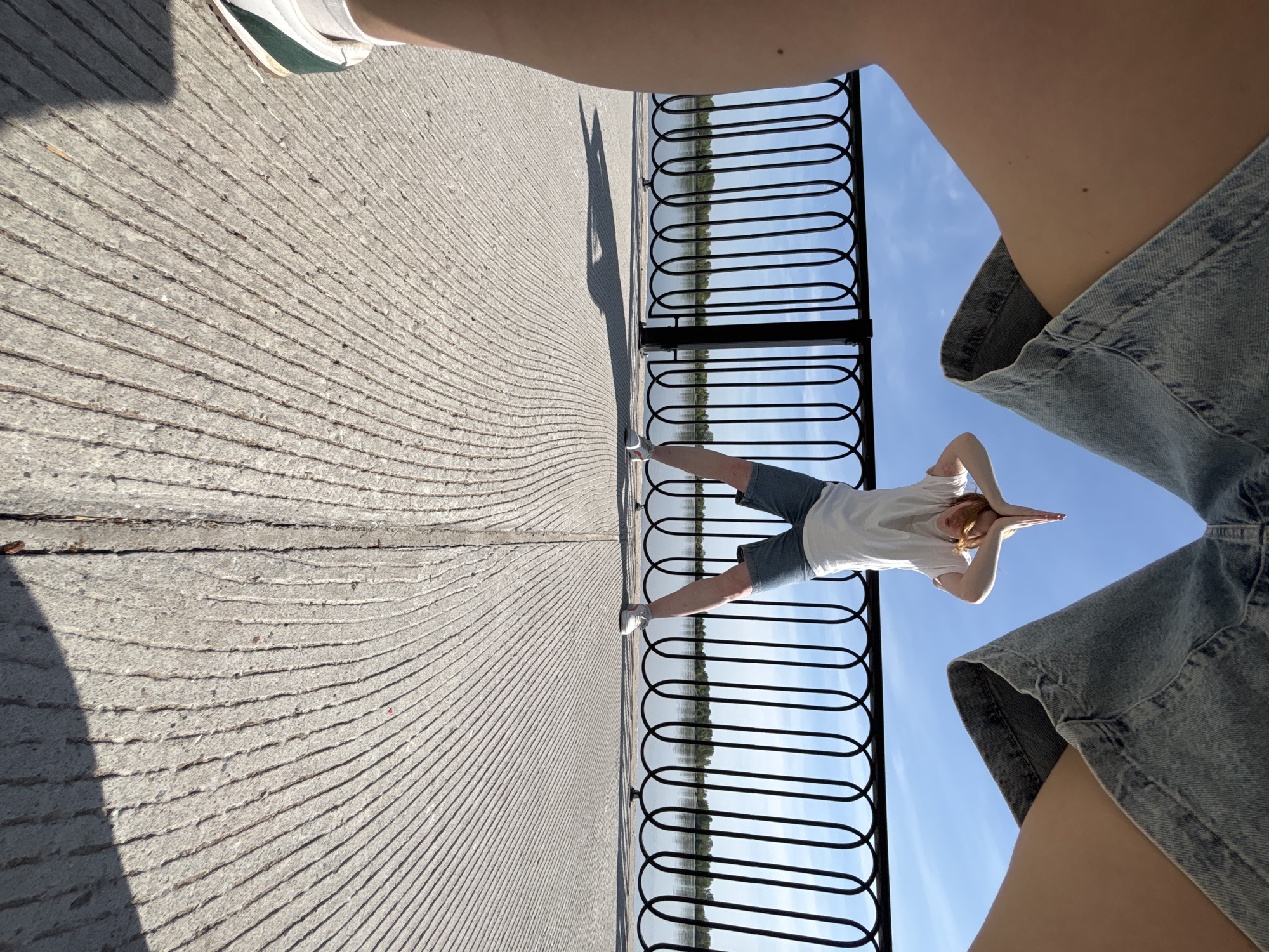 A person is photographed from below at a slightly tilted angle, looking upwards with the camera pointed at the sky. The foreground is dominated by denim shorts and a partial view of legs. A section of a black metal railing runs horizontally across the middle of the frame. Beyond the railing, there's a view of water and foliage, suggesting a river or lake. In the center, a person stands on a concrete surface making a triangular shape with their hands above their head. The concrete has a ridged texture. The lighting suggests it is daytime. The photograph was likely taken in the city of Perm, Russia.