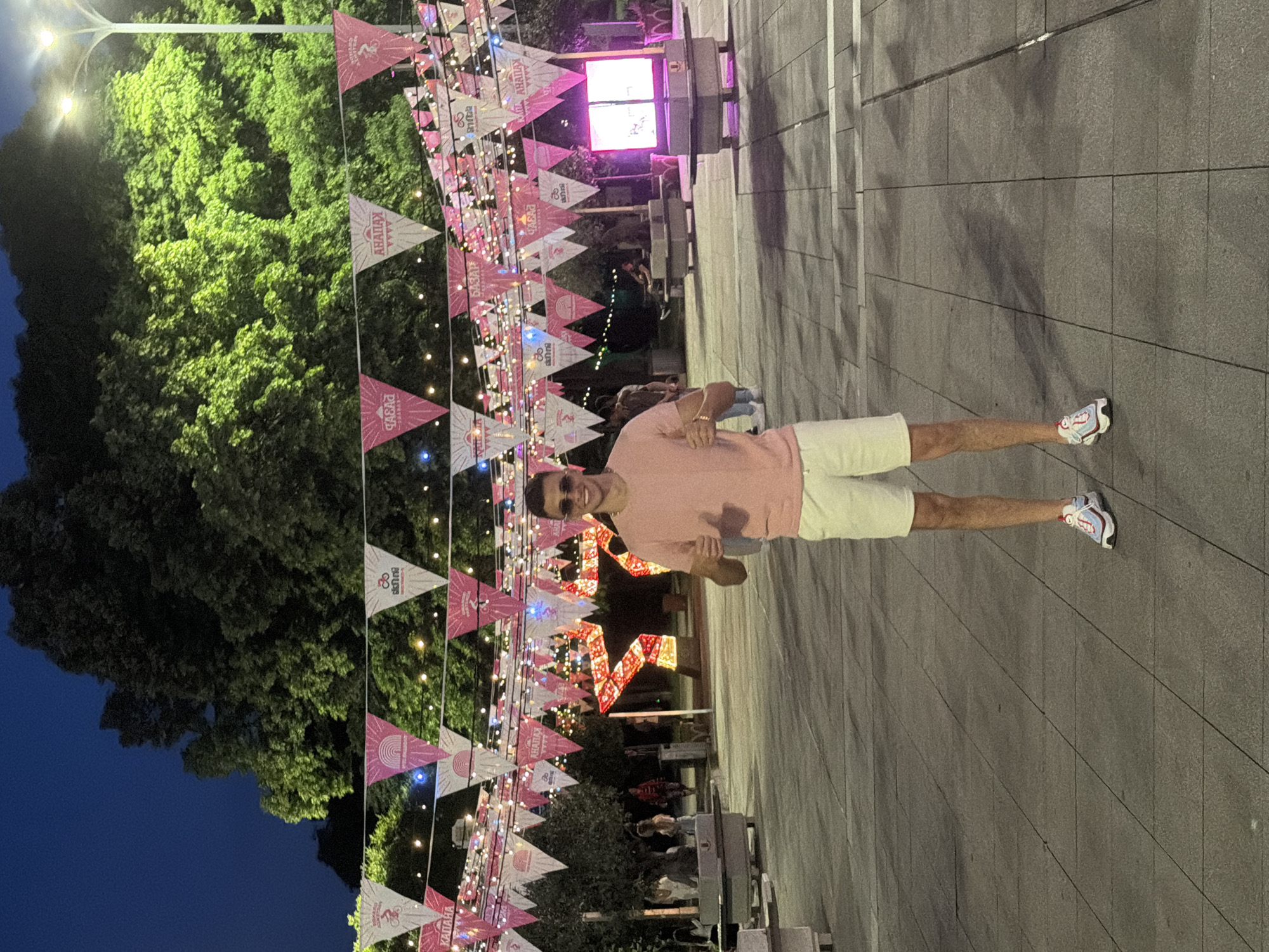 A man wearing a pink shirt, white shorts, and sunglasses stands on a paved city square at night. Pennant banners with logos or words are strung above the square, illuminated by string lights. A large, lighted, five-pointed star hangs in the background. Trees stand to the left, and a building with a brightly lit screen is in the distance. People are in the background, and benches are scattered on the right side of the frame.