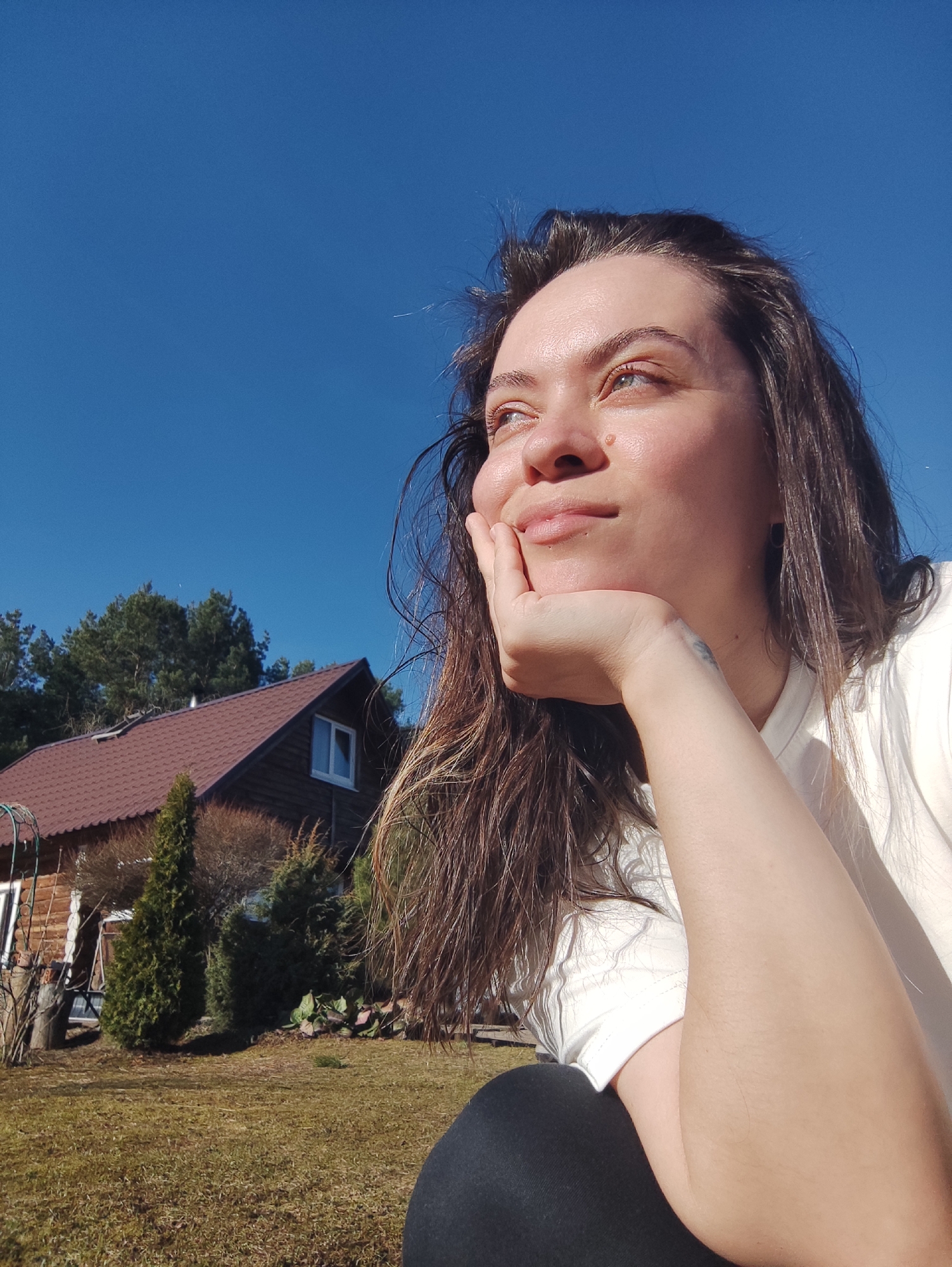 A bright outdoor photograph captures a young woman from a low angle, gazing upwards and to the left with a serene and contemplative expression, highlighted by a gentle smile. Her left hand rests under her chin, supporting her face. She has long, dark, slightly wavy hair and is wearing a light-colored t-shirt and dark leggings. A small, illegible tattoo is visible on her left wrist.
The backdrop consists of a clear, vibrant blue sky that dominates the upper half of the image. To the left, a rustic, dark brown wooden house with a reddish-brown shingled roof is visible, featuring a white-framed window. Several evergreen trees and bare bushes surround the house, with taller pine trees in the distant background. The foreground and midground feature patchy, somewhat dry grass.
The scene is bathed in natural sunlight, indicating a clear, sunny day, likely late morning or early afternoon. The overall atmosphere conveys peace and relaxation. This rural or countryside setting aligns with the provided location context of Фоки, Russia.