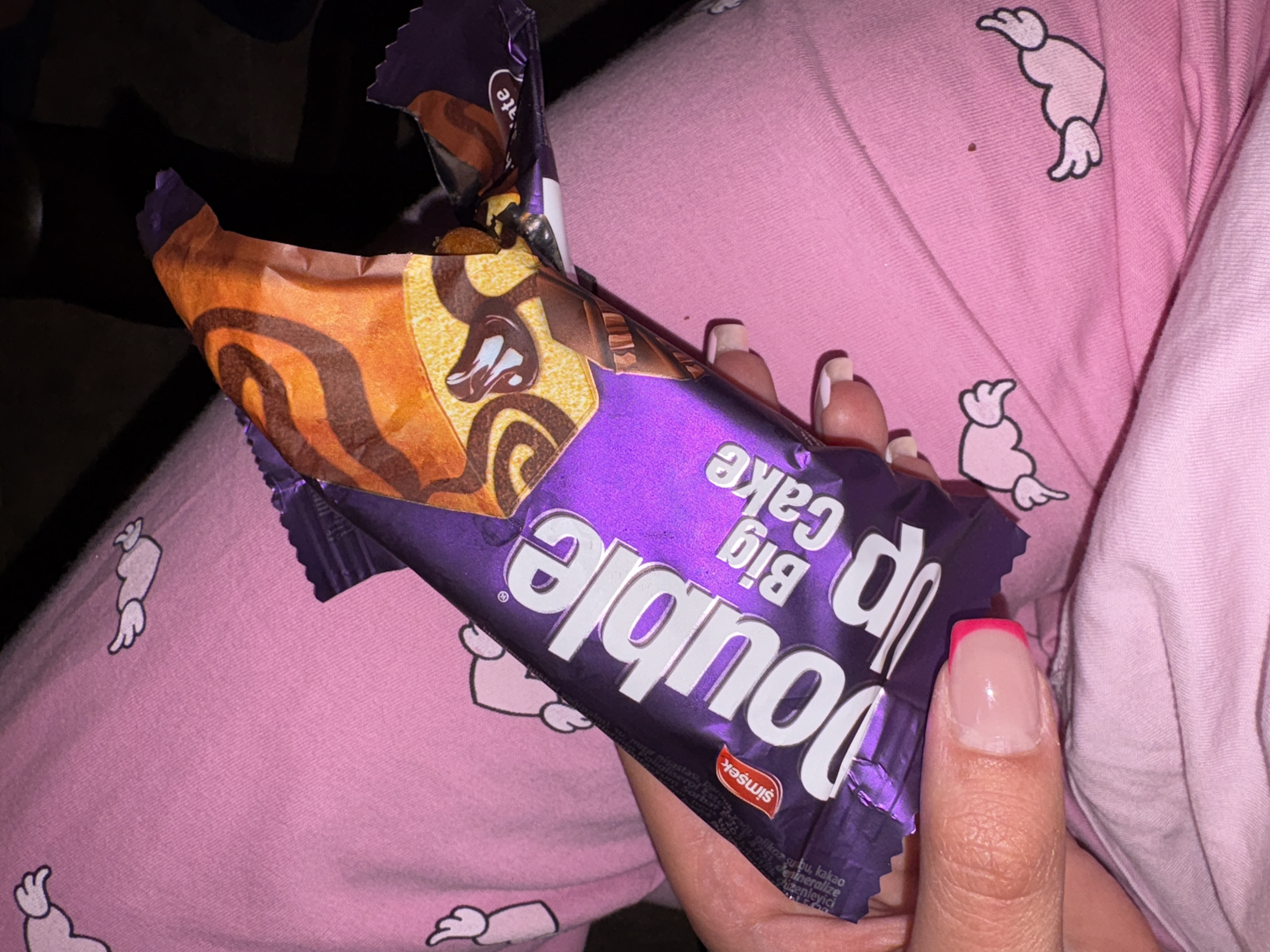 The image shows a close-up of a person's hand holding an opened snack bar or cake. The setting appears to be indoors, likely at night or in low light, suggested by the dark background. The hand is holding a purple and orange-colored wrapper for a product named "Double Up Big Cake," from the brand "Şimşek." The wrapper is partially torn open, revealing the cake inside, which has a swirled pattern of light and dark brown, indicative of vanilla and chocolate or similar flavors.
The person's hand has manicured nails with a French tip style, featuring a vibrant pink line at the tip over a natural, clear base. The hand is resting against a soft, pink fabric, which appears to be clothing or bedding. This fabric is patterned with repeated light pink or white cartoon-like figures that resemble abstract shapes or perhaps characters with wings/limbs.
Visible text on the packaging includes "Double Up Big Cake" and the brand name "Şimşek." Other partial words from the OCR, such as "nilkaz su ou, kakao," "denineralize," and "dizenleyici," suggest ingredients or product information, likely in Turkish. The activity shown is simply holding the snack, possibly about to consume it. The scene conveys a casual, relaxed moment. The location context provided is Vratsa, Bulgaria.