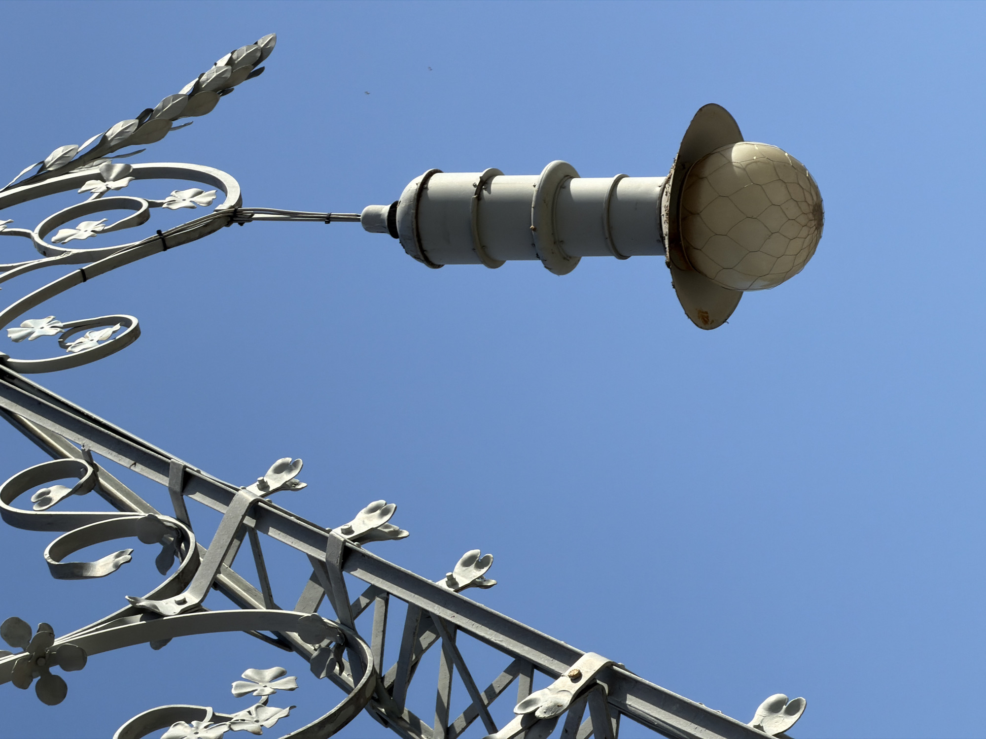 The image shows a close-up of an ornate, grey metal lamppost against a clear blue sky. The lamppost features decorative elements resembling flowers and leaves, attached to a lattice-like structure. A grey, cylindrical lamp housing with several ribbed sections hangs from the structure, topped by a conical element. Below the housing is a spherical lamp, enclosed in a wire mesh, with a flared, rusted metal base. The image appears to be taken outdoors, possibly in a public space given the nature of the lamppost. The lamppost's intricate design suggests it could be a feature of a historic or architecturally significant area. The clear sky implies fair weather. The image does not contain any people or actions. The style of the lamppost is reminiscent of early 20th-century Parisian or Barcelona street furniture.
