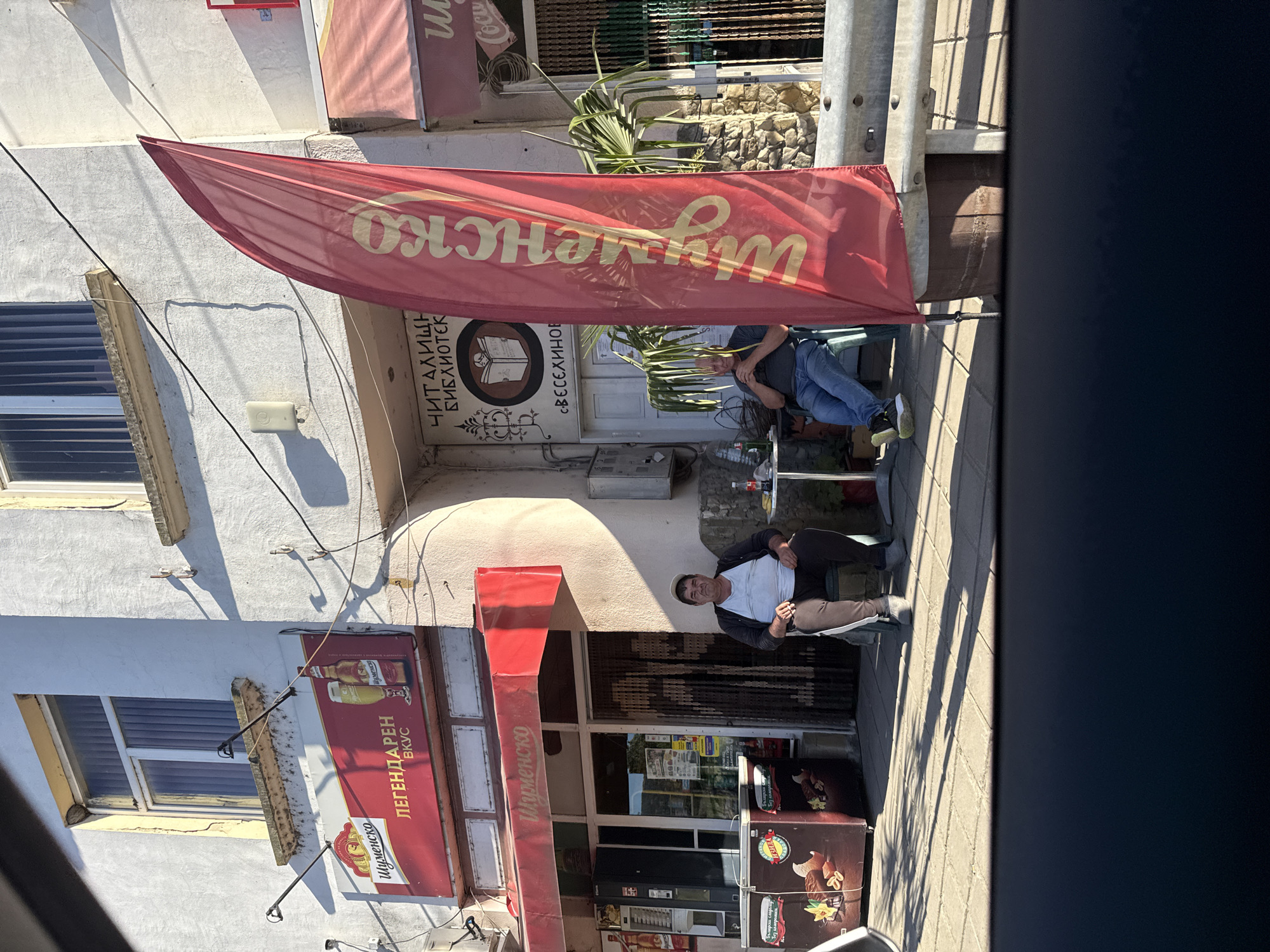 This image depicts the exterior of a commercial establishment in the city of Veselinovo, Bulgaria. In the foreground, a large red flag with yellow lettering, likely advertising a brand, stands prominently. Two men are seated on chairs outside the building. The man on the left is wearing a light-colored t-shirt, dark pants with a white stripe, and a dark jacket. He is looking towards the camera. The man on the right is wearing a dark t-shirt and blue jeans, and is also seated. In front of the building, there is an ice cream freezer. Above the entrance, a sign displays text in Cyrillic, and to the left of that, another sign features a beer bottle and glass. The building itself appears to be painted white and has multiple windows. The ground is paved with interlocking bricks. The overall scene suggests a casual daytime setting outside a shop or cafe.