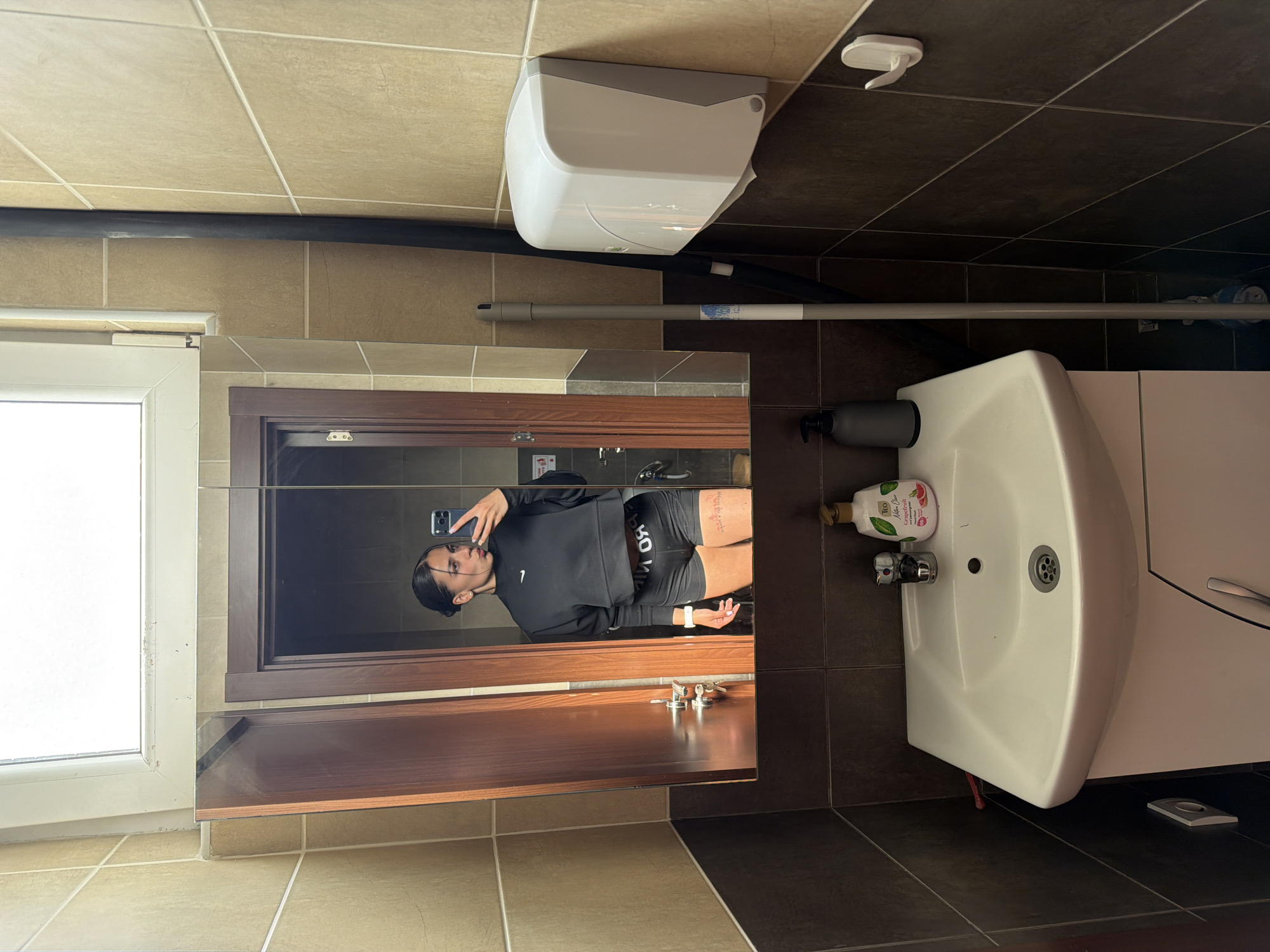 The image depicts a bathroom scene, likely in a residential setting. A young woman is reflected in the mirror, holding a smartphone, apparently taking a selfie. The bathroom features light brown wall tiles and dark brown floor tiles. A white hand dryer is mounted on the wall. Below the mirror, a wooden shelf is visible. A white sink and vanity are positioned on the right side of the image. Various toiletries such as soap and lotion are placed on the sink. A mop is leaning against the wall above the sink. A window is visible on the left side of the mirror. The city of Gabrovo, Bulgaria is not discernible from the visual information in the image.