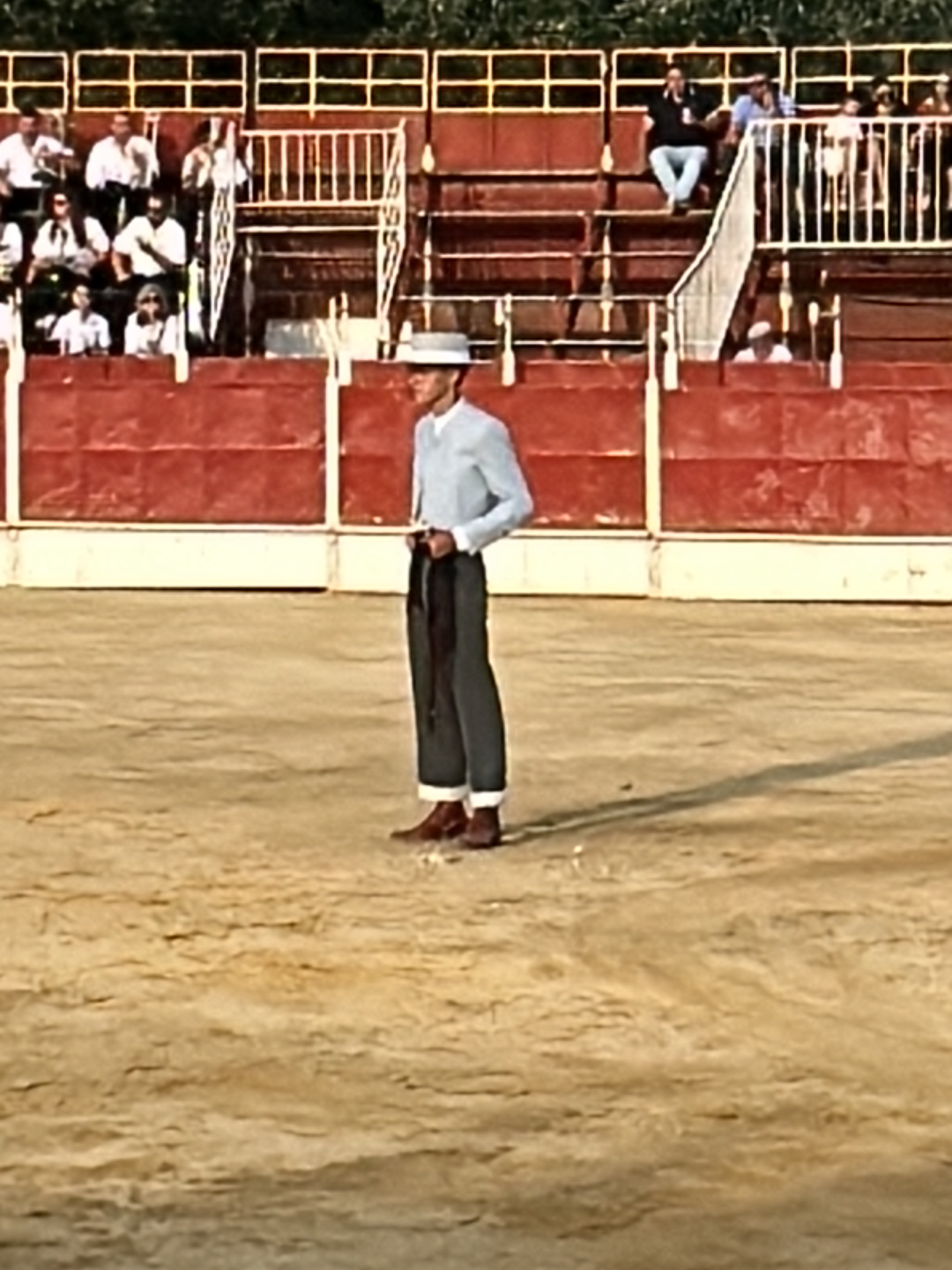 A man in a light-blue jacket and white hat stands in a bullring in San José del Valle, Spain. The bullring has a red barrier and stands for spectators in the background. The sand of the bullring is light brown. The man is wearing dark trousers and brown shoes.