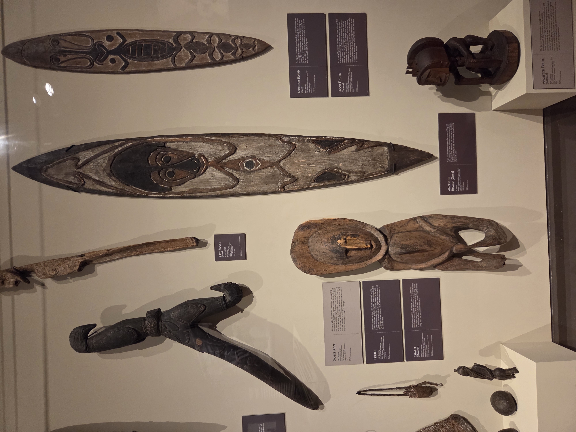 The image displays a museum exhibition of Oceanic artifacts. In the center foreground, a wooden ancestral figure with detailed carvings stands on a white pedestal. To its left, a smaller, dark wooden figure is presented alongside a smooth, dark, ovoid object and a woven pouch with a handle. Above these, a long, dark, intricately carved wooden object with a hook-like element is displayed horizontally. Further up the wall, a large, elongated wooden shield with painted designs is mounted vertically. To its right, another, narrower wooden shield with similar painted motifs is visible. Smaller display cards with text are positioned near some of the artifacts. The overall environment suggests an indoor museum setting with controlled lighting. The exhibition is located in Santa Ana, United States of America.