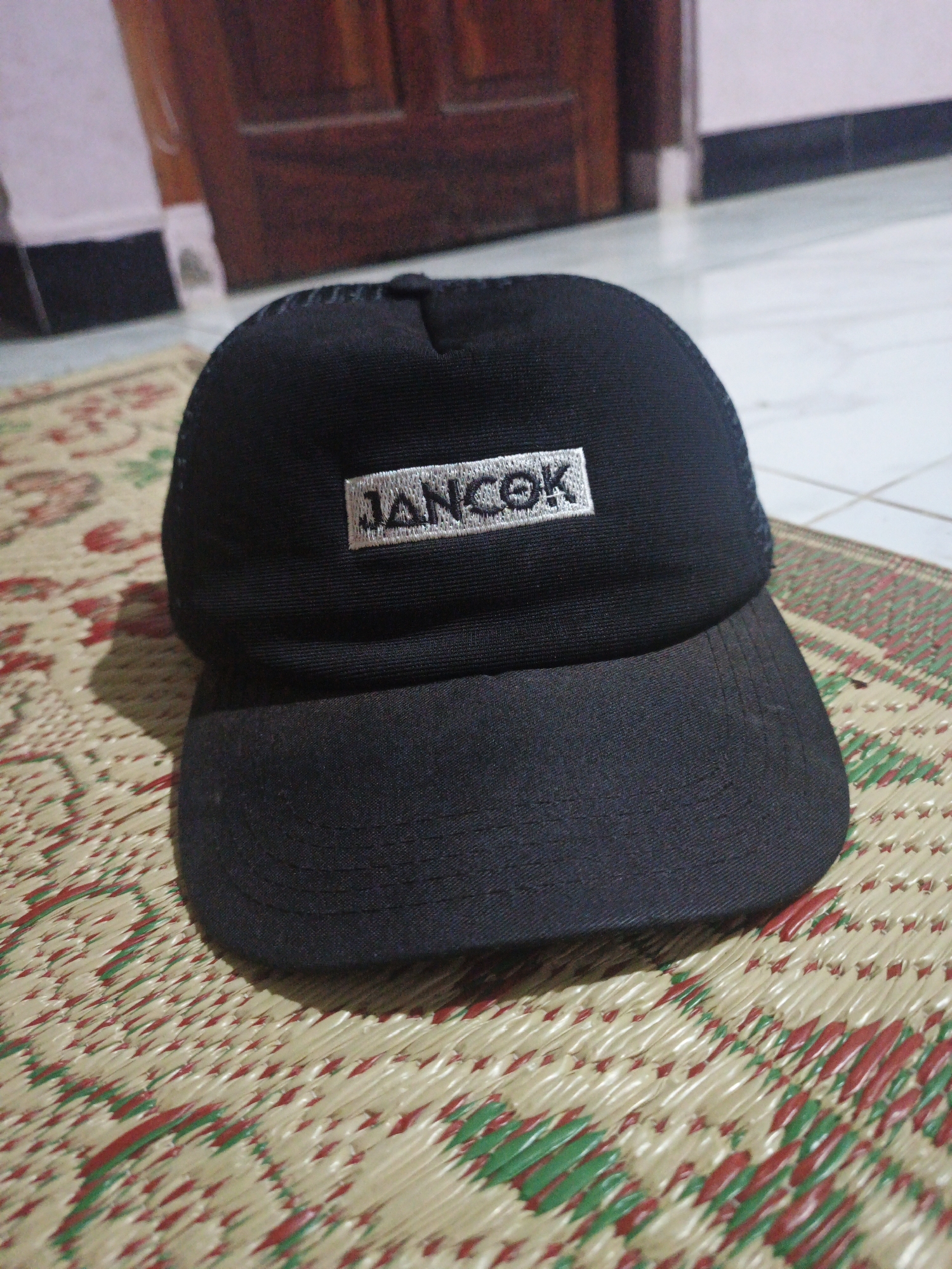 A black baseball cap with "JANCOK" embroidered in white on a rectangular patch is placed on a woven mat. The cap has a mesh back and a flat brim. The mat has a pattern of alternating red and green woven threads on a cream background. In the background, a wooden door and a tiled floor are visible. The scene appears to be indoors.