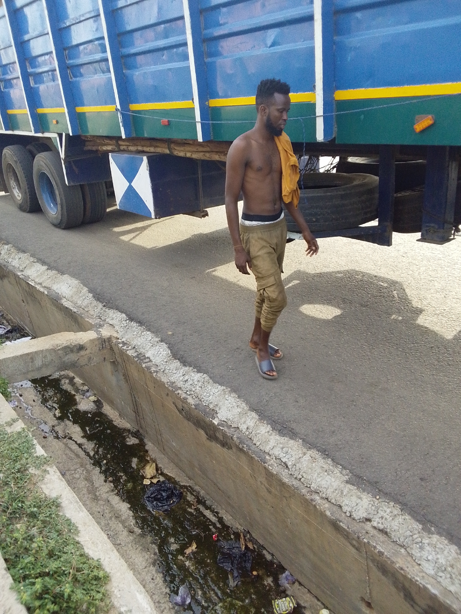A shirtless man is walking along a road beside a large blue truck in Lokoja, Nigeria. He is wearing tan pants and sandals, with a yellow cloth draped over his shoulder. The truck has a yellow stripe along the bottom and visible tires. Next to the road is a concrete drainage ditch filled with debris. The asphalt road appears to be sunny and dry.