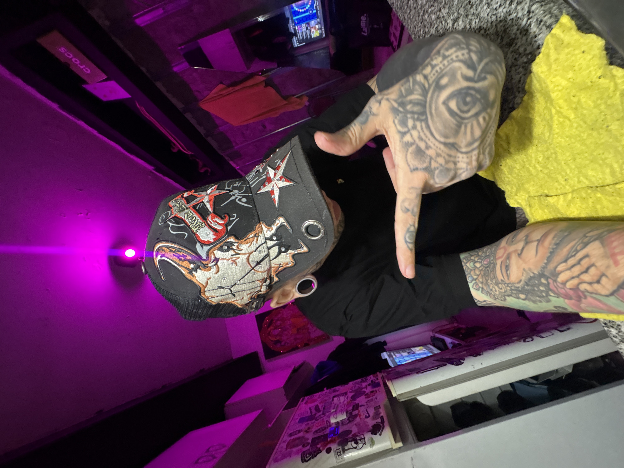 An individual is captured indoors under magenta lighting. The subject wears a black t-shirt, a black trucker cap with embroidered skull and star motifs, and large ear gauges. Extensive tattoos cover both forearms, including a figure resembling a deity on the right forearm, and intricate patterns across the right hand, with solid black tattooing on the palm side of the fingers. The subject's right hand is raised, index finger extended left, and thumb upright. A yellow, textured material is visible below the right hand. The background includes a shelf with a 'crocs' label, a sticker-covered surface, artwork displaying a glowing red object, and a computer monitor showing a colorful interface next to a 'Laime.' branded bag. A red fabric item hangs further back. The precise location within Buenos Aires, Argentina, is not ascertainable from the image.