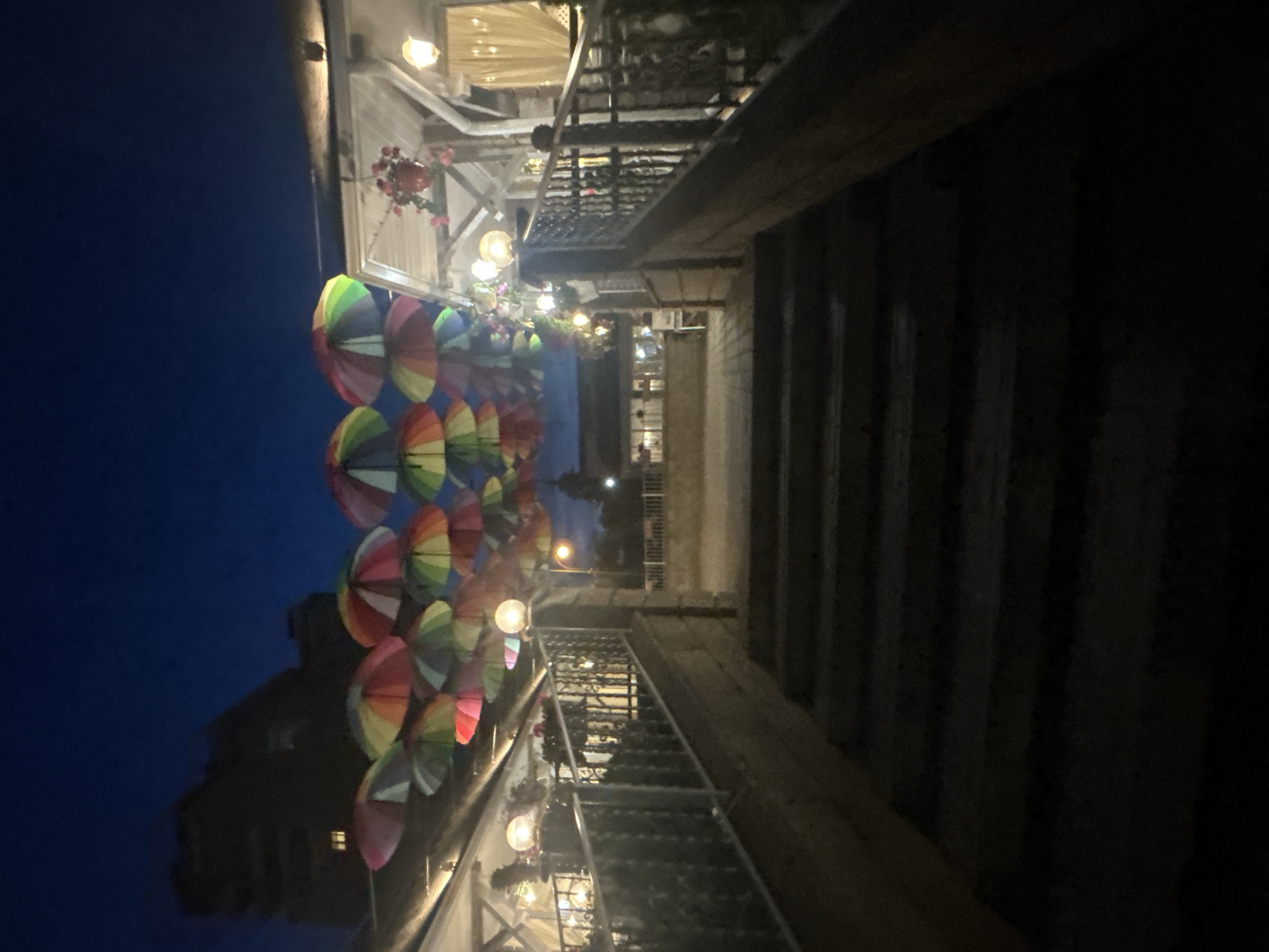The image depicts an outdoor staircase leading up to a dimly lit, open-air establishment at dusk. Above, a cluster of colorful umbrellas are suspended, creating a decorative canopy. The surrounding environment suggests a commercial area, possibly a cafe or restaurant, given the outdoor seating and lighting. In the background, a silhouetted building rises against the dark blue sky. The overall scene is characteristic of an evening ambiance in a public space, possibly in the city of Brest, Belarus, known for its historical sites and recreational areas that often feature such decorative elements.
