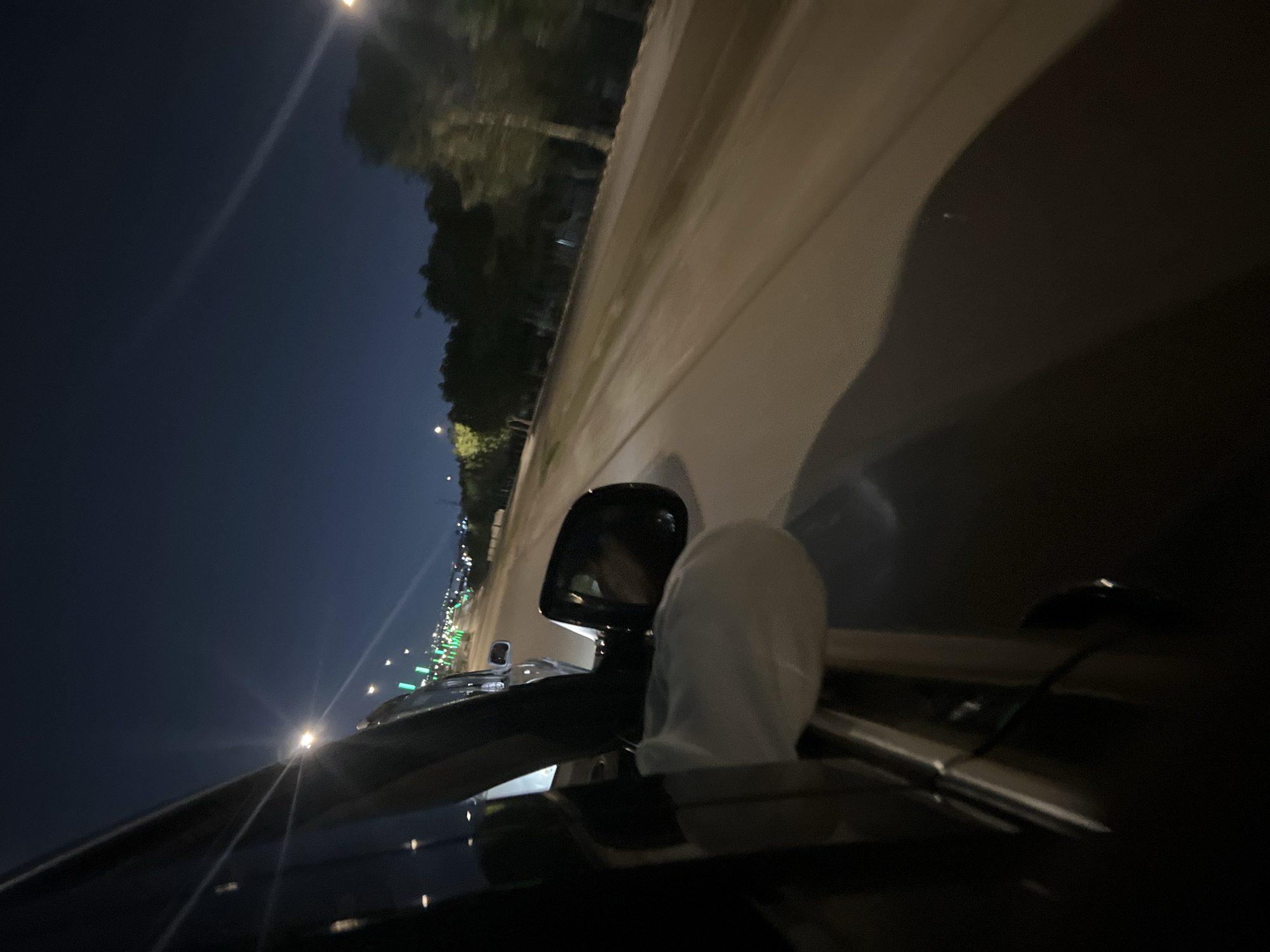 The image depicts an eye-level perspective from inside a moving car at night. The car is on a road in Abuja, Nigeria. On the left, trees and street lights line the road under a dark sky. On the right, there is a blurry view of the side of the road. The rearview mirror is visible, reflecting the driver's face. A white cloth or object is draped over the car's interior, partially obscuring the view. The car's dashboard and windshield are visible in the foreground.
