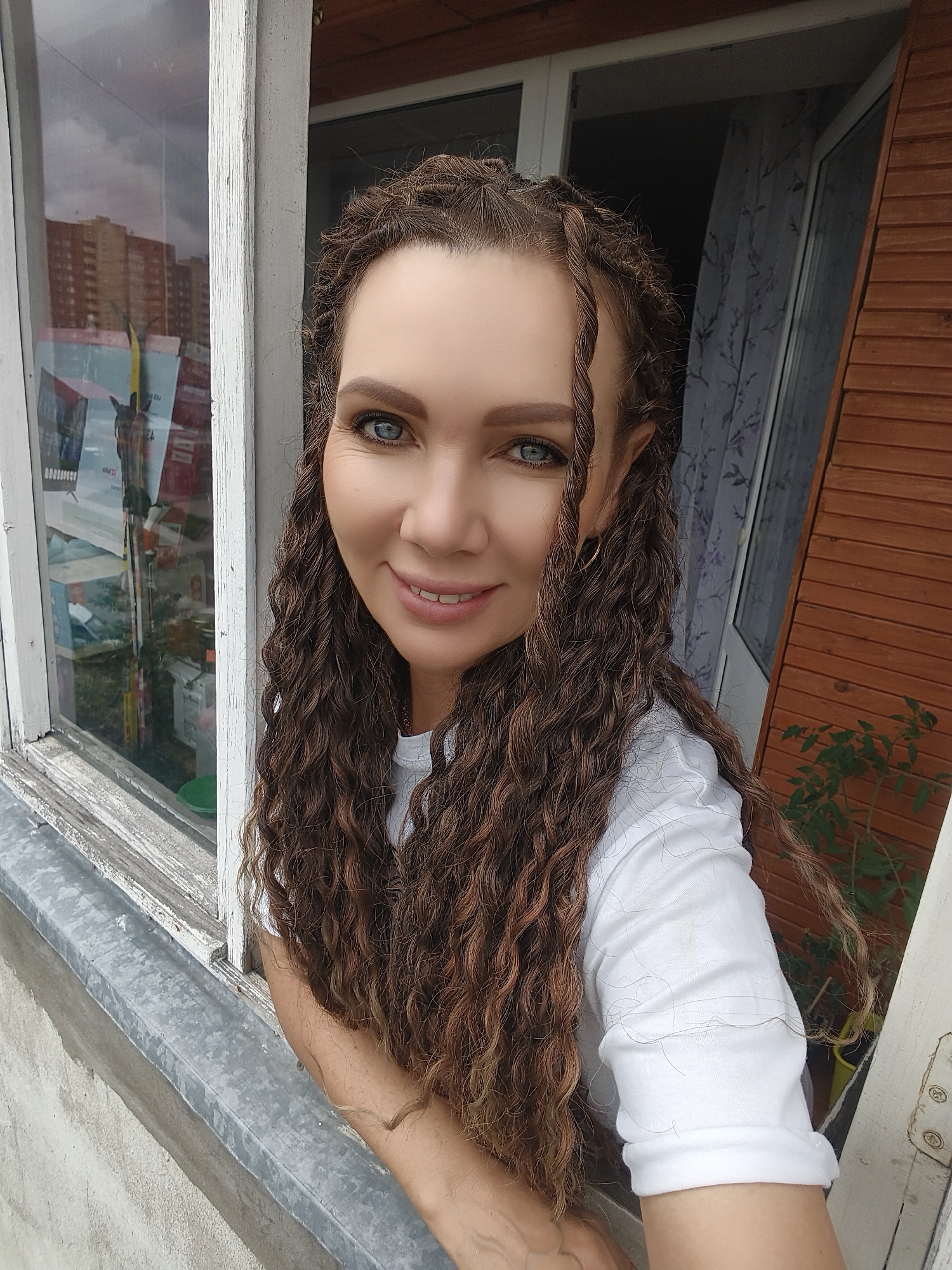 A medium-complexioned woman with blue eyes, light brown eyebrows, and a slight smile is depicted in a medium-close-up, looking directly at the viewer. Her long, dark brown hair features numerous dreadlocks or tight braids, with a few strands pulled forward around her face. She wears a plain white short-sleeved t-shirt, and a small gold hoop earring is visible in her right ear.
She is positioned at an old, weathered white window frame, leaning slightly into or out of it, with her right arm visible on the metal windowsill. The exterior of the building appears to be concrete or stucco below the sill.
Through the window on the left, reflections show multi-story buildings under a cloudy sky, consistent with an urban environment. Blurred shapes and colors suggest advertisements or posters are also reflected in the glass. To the right of the woman, beyond the window frame, an interior space is visible, featuring a brown wooden slatted wall, a white door or additional window frame, and a curtain with a purple floral pattern. A small potted plant with green leaves is visible at the bottom right, suggesting a domestic setting. The exact city and country are unknown.