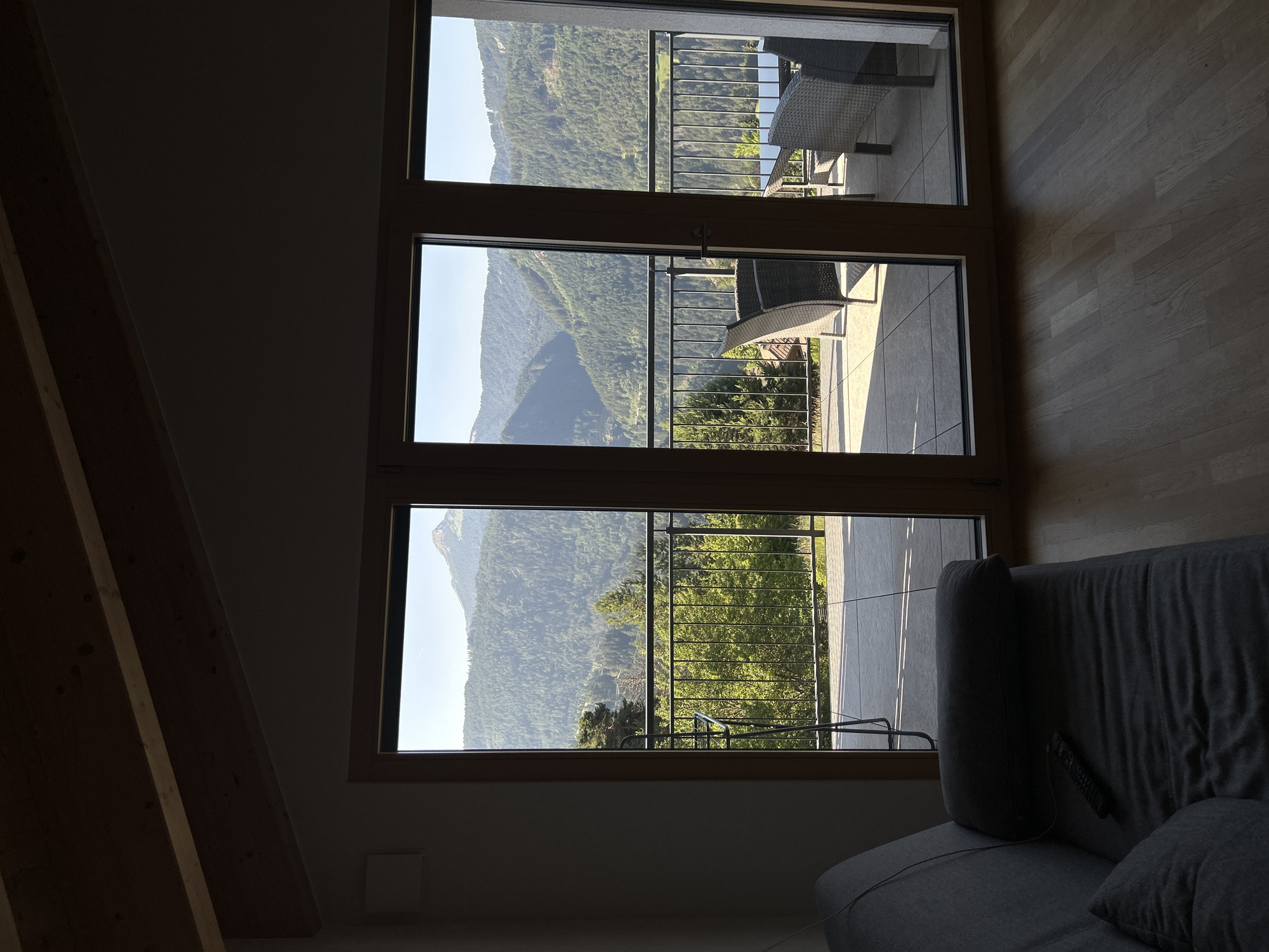 A wide shot captures an interior living space opening onto a balcony with a mountain view. In the foreground, a dark gray sofa is positioned on the left, with a remote control resting on its surface. The floor is made of light-colored wood planks. The room features a sloped ceiling with exposed wooden beams. Large glass doors lead to a balcony. Outside, a metal railing lines the balcony, beyond which a picturesque mountain range covered in dense green trees stretches across the horizon under a clear blue sky. The balcony appears to be furnished with outdoor seating, including wicker chairs and a table. The lighting suggests it is daytime, with sunlight casting shadows.