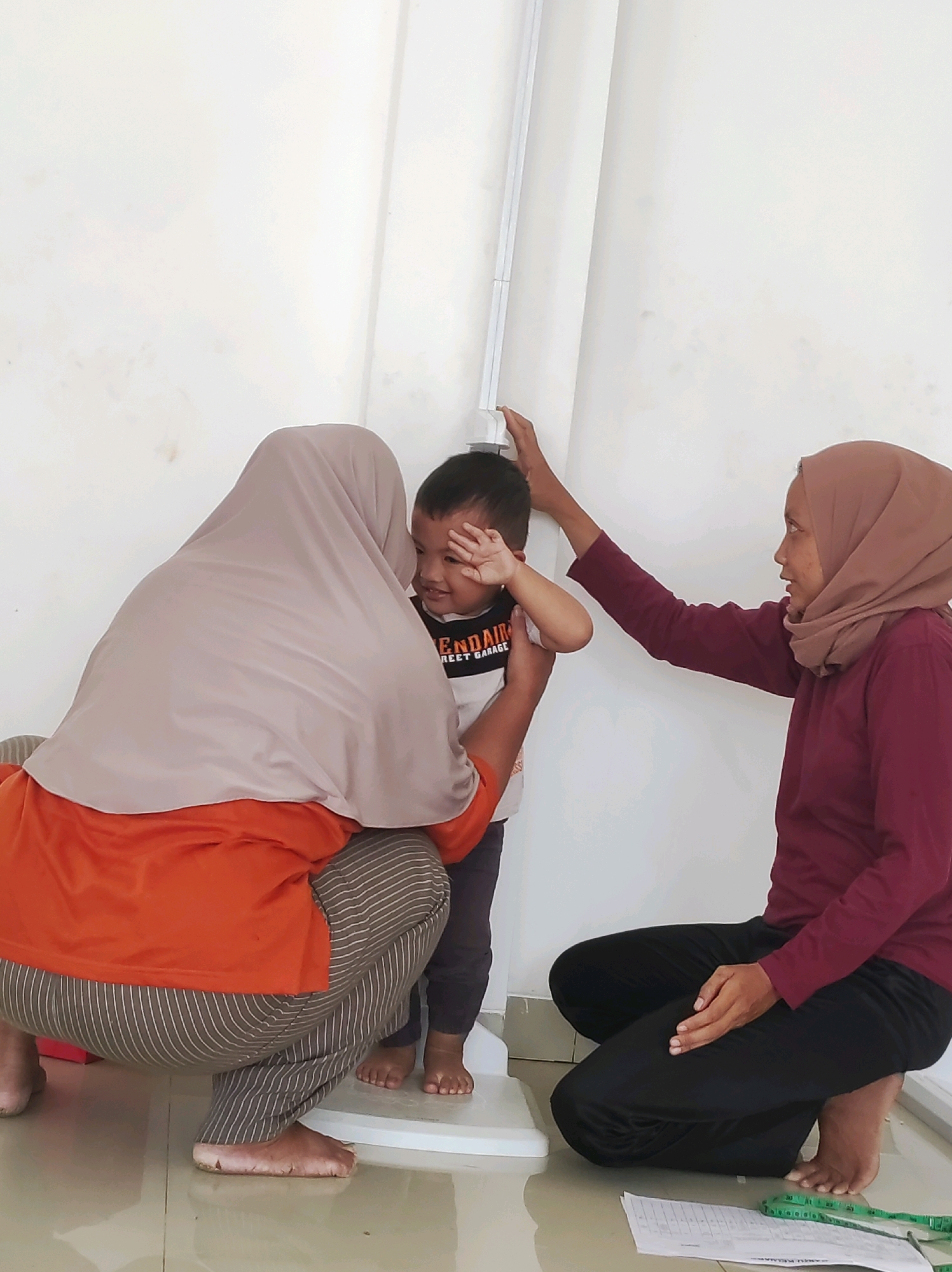 The image depicts a scene in Mojopuro, Indonesia, where two women are assisting a young boy in being measured for height. One woman, wearing a light brown hijab and an orange shirt, is crouched down and appears to be holding the boy's feet. The other woman, with a darker brown hijab and a maroon long-sleeved shirt, is standing and using a measuring device attached to the wall to determine the boy's height. The boy, wearing a white t-shirt with black and red lettering, is standing on a white scale, his left hand covering his eyes. The environment appears to be indoors, with a plain white wall and a tiled floor. A piece of paper and a green measuring tape are visible on the floor in the foreground.