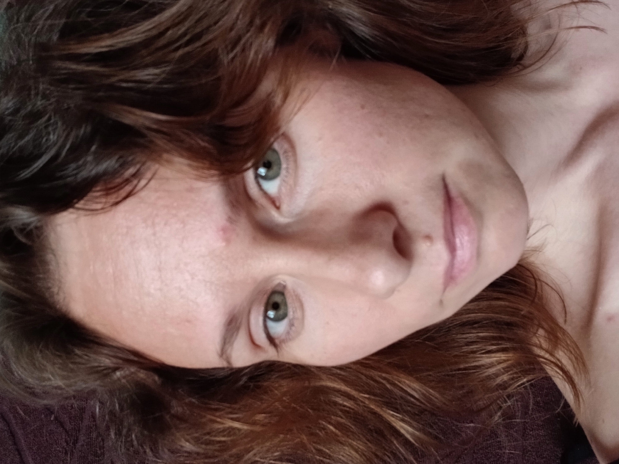 A close-up, high-angle shot captures the face of a woman with medium-length, wavy brown hair. Her eyes are green and her skin is fair with visible pores and a few freckles. She has a small mole on her upper lip and a red blemish on her forehead. The background is a dark, textured fabric. The image does not contain information that could place it in Yekaterinburg, Russia.