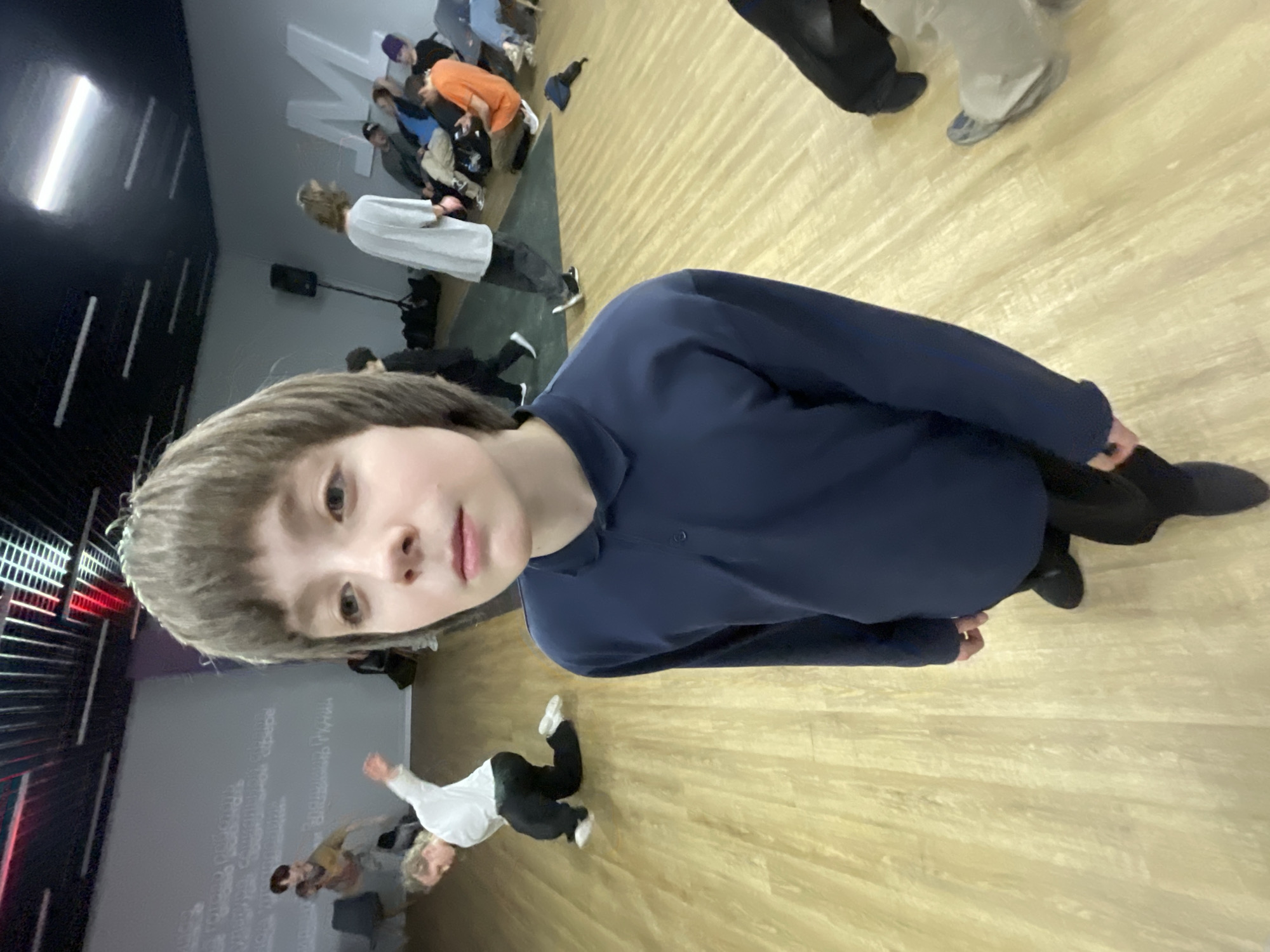 The image is a vertical selfie taken indoors, showing a young person in the foreground and a busy studio environment behind them.
The primary subject is a young person with light brown, shaggy hair and bangs, looking directly into the camera with a neutral expression. They are wearing a dark blue long-sleeved shirt and what appear to be dark pants or leggings, with a glimpse of dark shoes. Their head and upper torso dominate the upper half of the frame.
The background reveals an active, brightly lit indoor space, likely a dance or rehearsal studio. The floor is made of light-colored wood with a noticeable grain pattern. On the left side of the image, there's a dark wall with several horizontal bright light strips, and one prominent vertical light fixture. Adjacent to this, there's a light gray wall or screen with visible writing. Large white Cyrillic letters, possibly "MAM" or "ПЛАН", are prominent on this surface. Below this, there are lines of handwritten or projected text, also in Cyrillic, with some words partially legible, such as "Музыка" (Music).
Several other people are scattered throughout the background, engaged in various activities. In the mid-ground, a person wearing a white long-sleeved shirt and dark pants is performing a dynamic pose or movement on the wooden floor, their body angled, and hands near the floor. Further back, near the gray wall, a group of people is visible. One individual wears an orange shirt and appears to be sitting or leaning forward. Another person in this group has a purple beanie. Other individuals are standing or sitting, dressed in casual attire. A black speaker on a stand is also visible in the mid-ground. Towards the far right, the lower legs and feet of additional individuals can be seen, some wearing light-colored shoes and pants.
The overall scene suggests a vibrant atmosphere, possibly during a dance class, rehearsal, or performance preparation, with participants either practicing or observing.