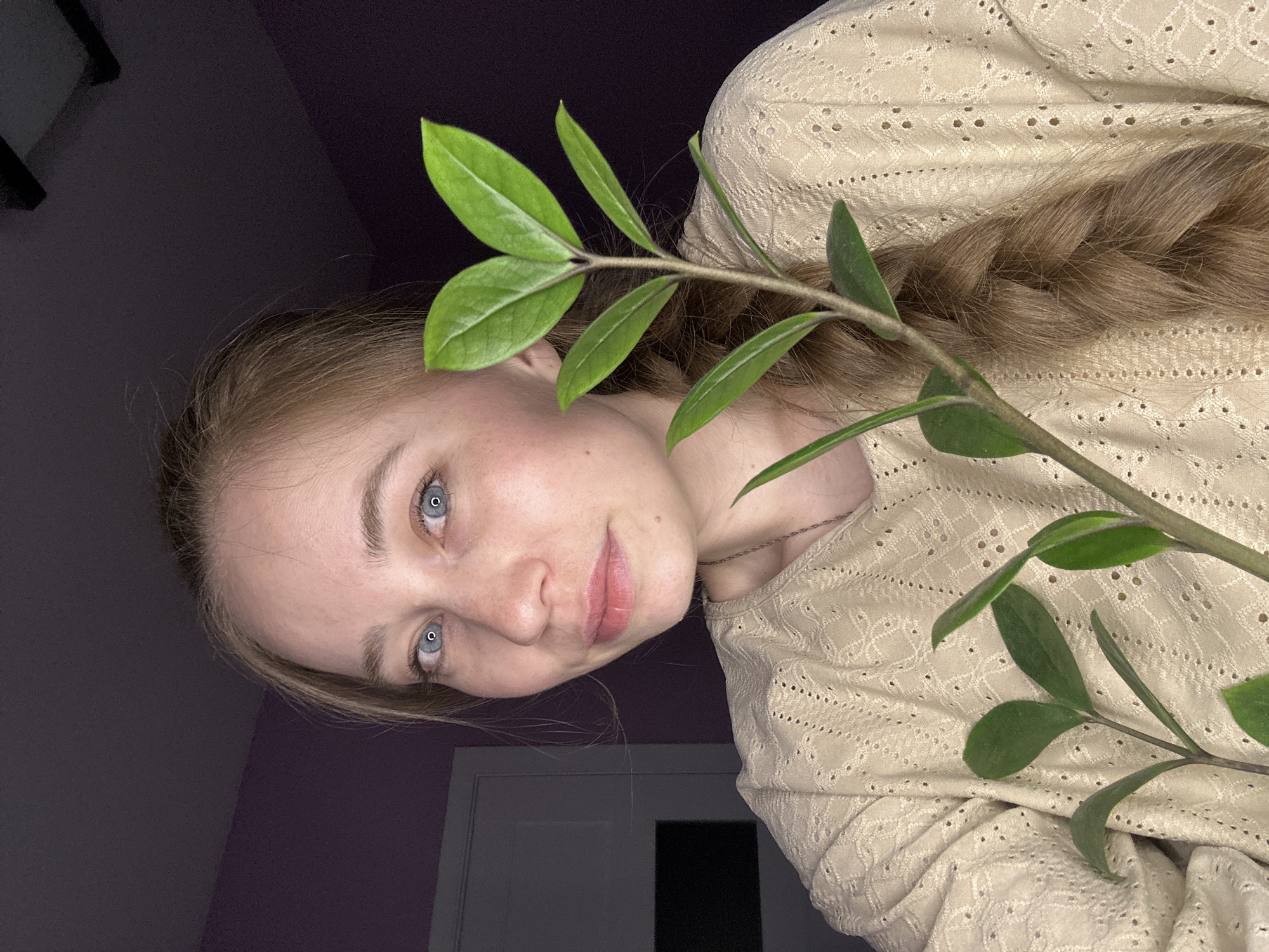 A young woman with fair skin and light blue eyes is posing with a leafy green plant. She is wearing a light beige-colored, long-sleeved top with a textured pattern. Her light brown hair is braided and falls across her shoulder. The background is a combination of dark and light purple walls, with a white door visible. The image is taken from a slightly low angle. There is no information to suggest that this picture was taken in Saint Petersburg, Russia.
