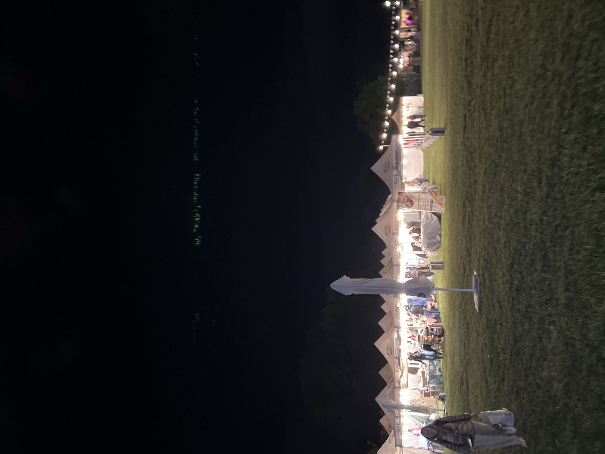 A nighttime outdoor art bazaar event is taking place in a grassy field. Several white tents with pointed roofs are set up in a row, illuminated from within. Signs on the tents read "Art Bazar". People are visible walking around and interacting within the tent areas. In the foreground, a woman with a light-colored jacket and pants walks to the left, carrying a tote bag. She appears to be walking away from the bazaar. The sky above is dark, with some faint lights visible in the distance. The scene suggests a community event or festival.
