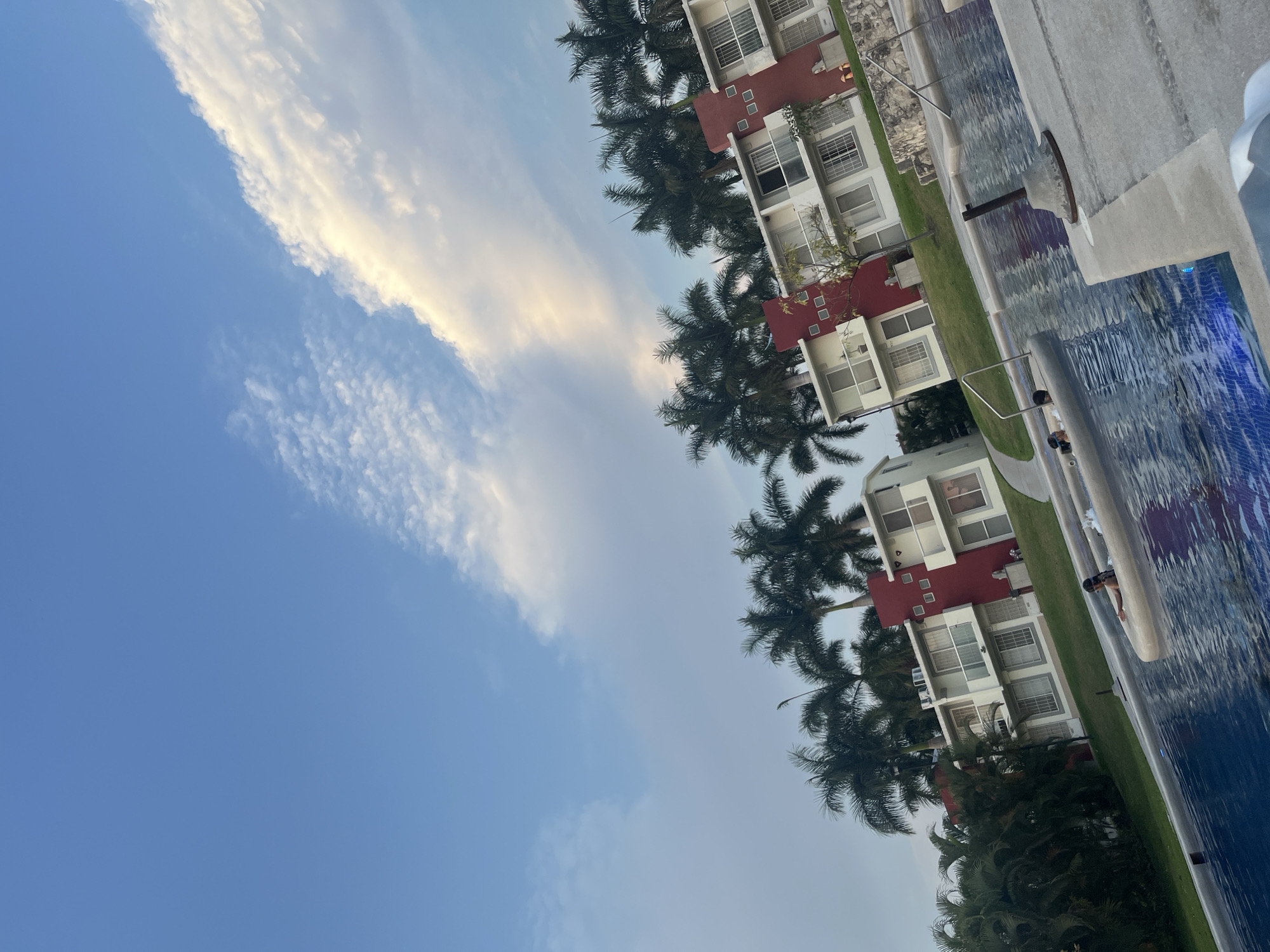 The image captures a scene in Xochitepec, Mexico, featuring a swimming pool adjacent to a set of buildings. Palm trees are interspersed between the structures. The pool's water reflects the sky, and the buildings appear to be residential, with multiple windows and a red architectural feature on top. A blue sky occupies a significant portion of the frame, with scattered clouds. A person is visible in the pool.