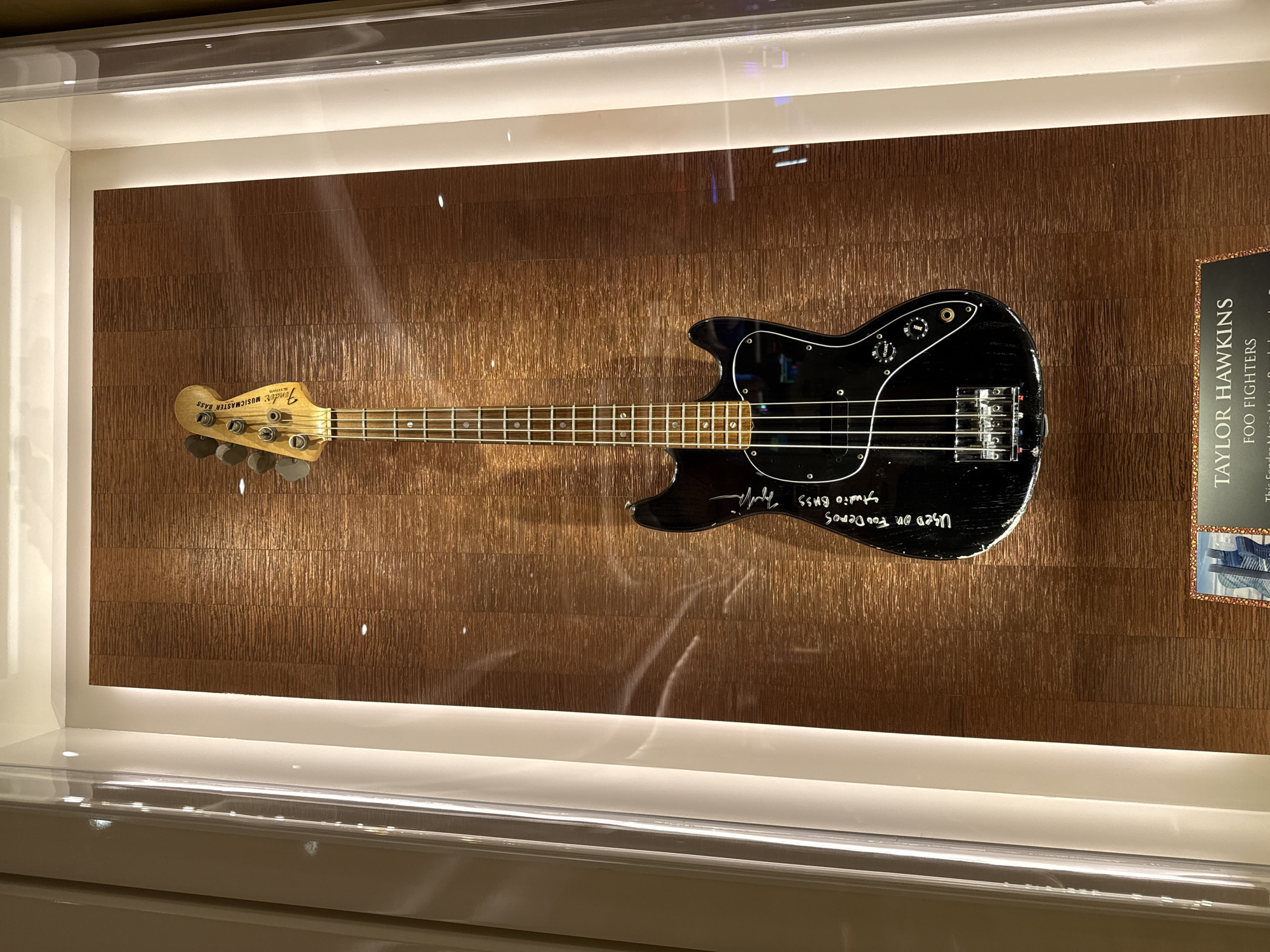 This is an indoor photograph of a black Fender Musicmaster bass guitar displayed in a glass case against a textured brown background. The guitar is positioned horizontally, with the headstock pointing left and the body pointing right. The headstock displays the Fender logo. White writing is visible on the body of the guitar, reading "Used on Foo Deoes Studio Bass." A sign on the right side of the case reads "Taylor Hawkins Foo Fighters." The display case is brightly lit. The photograph was taken at the city of Hollywood Seminole Reservation, United States of America.