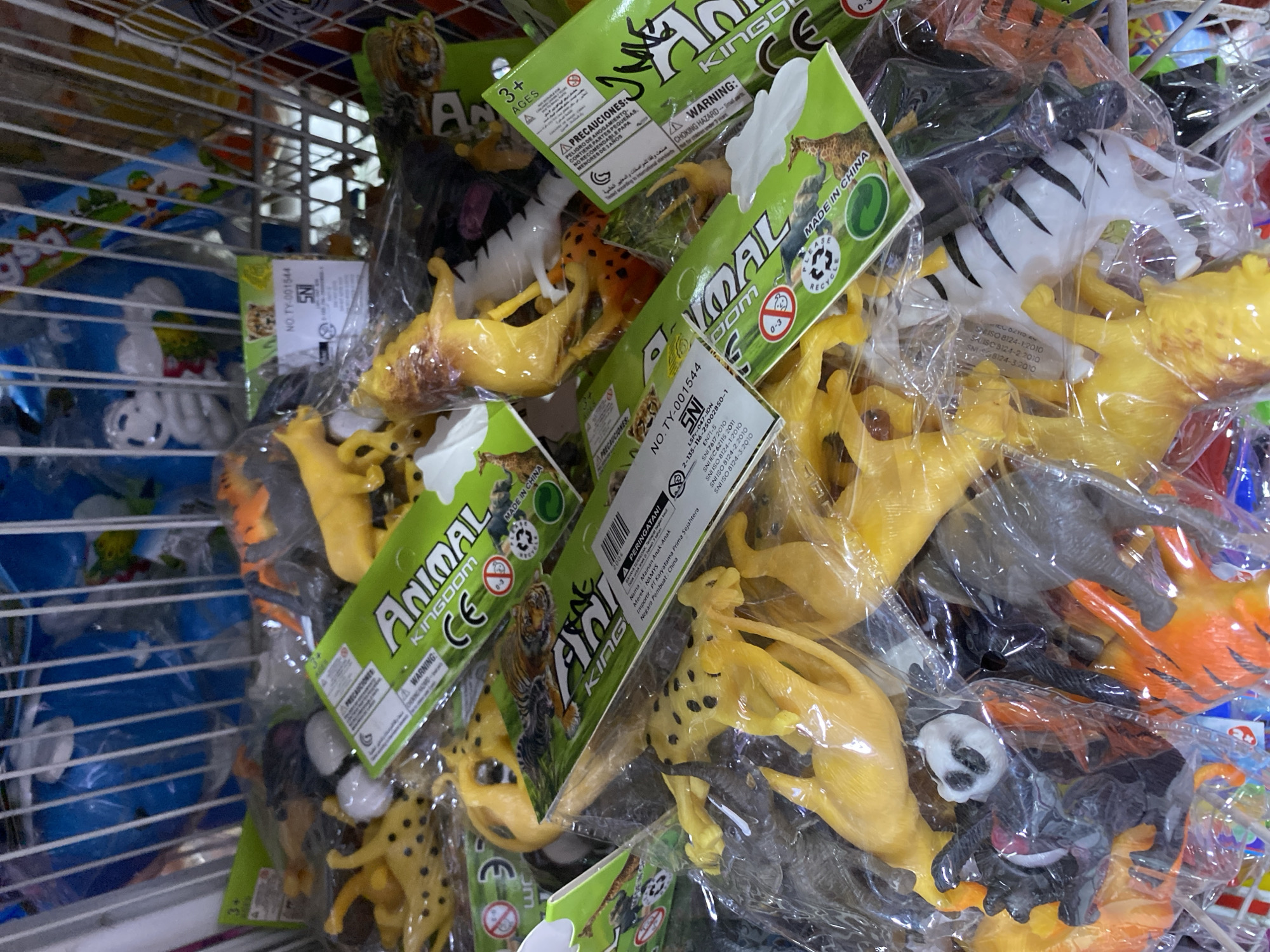 The image shows a close-up, overhead view of numerous small plastic animal toys in plastic packaging. The toys are various animals such as lions, tigers, cheetahs, zebras, and what appears to be a panda. They are primarily yellow, orange, black, and white. The packaging is predominantly bright green with text and images of animals. Some of the packaging has labels that read "Animal Kingdom," "Made in China," and warning symbols. The toys are crammed into a wire basket, suggesting they are for sale. In the background, there are blurred blue and white objects, possibly other toys or merchandise. This scene is consistent with a display of toys in a market stall or a small shop in Banda Aceh, Indonesia, where such items are commonly sold.