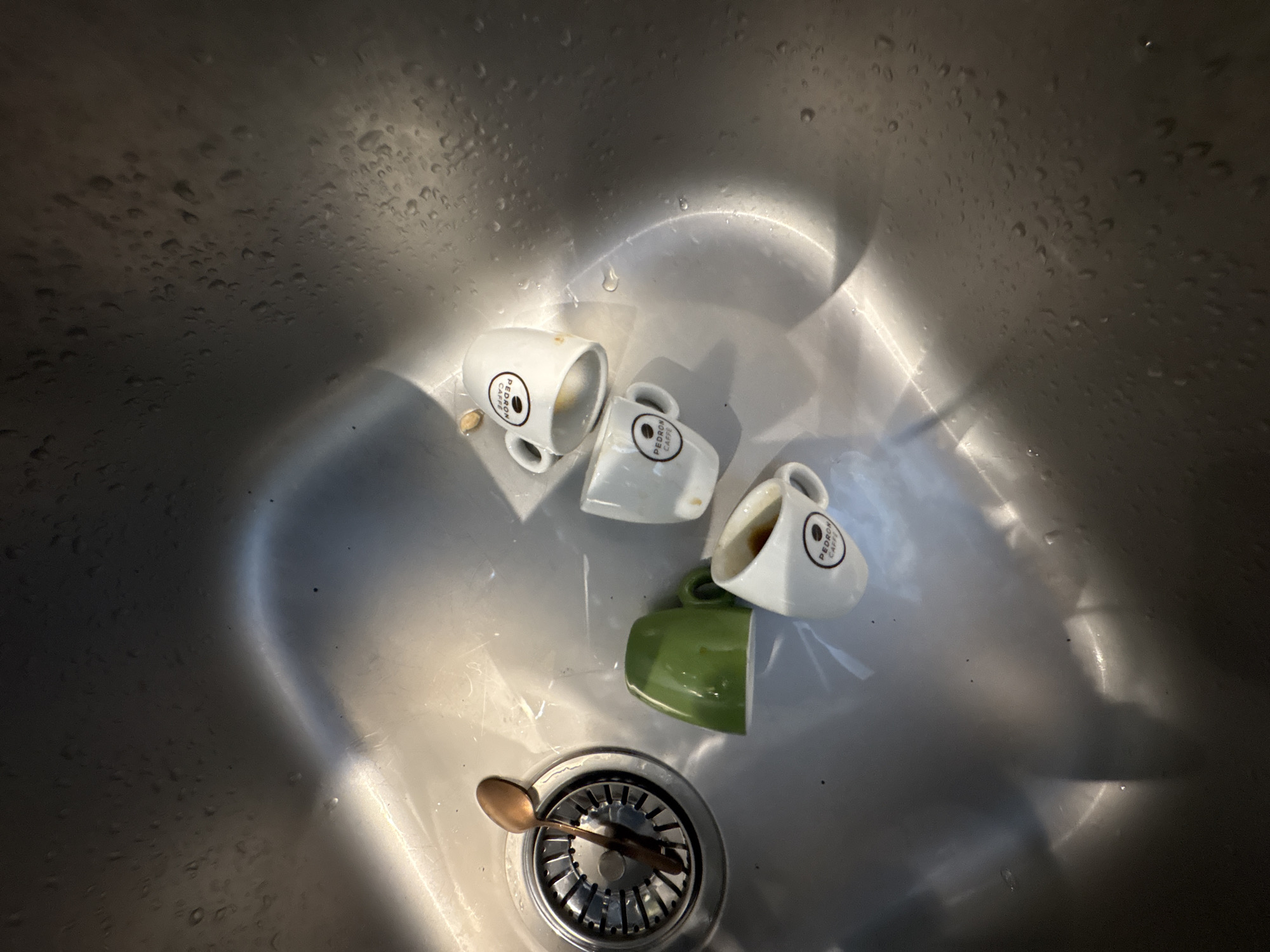 The image shows a stainless steel sink with water droplets on its surface. Inside the sink, there are four small coffee cups, three of which are white with a "PEDRON CAFFÈ" logo and one is green. A copper-colored spoon rests on the drain cover. The sink appears to be in a kitchen or utility area. There is no indication of a location in Sofia, Bulgaria.