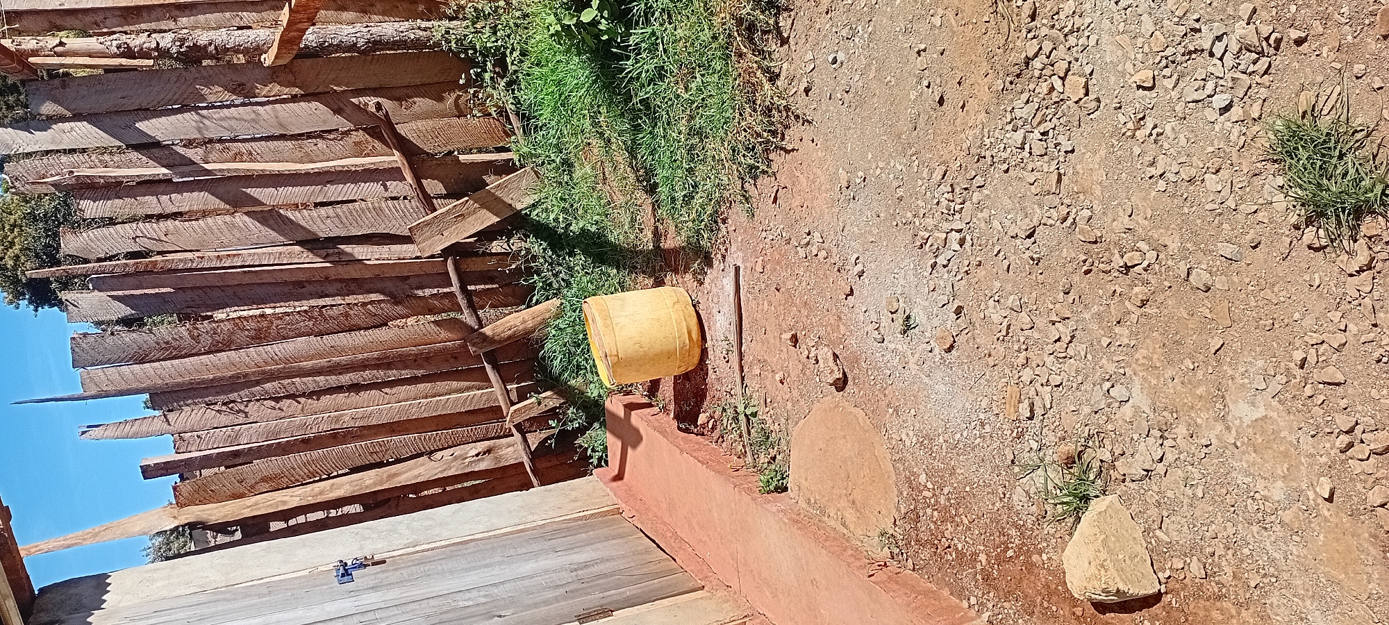 The image depicts an outdoor scene, likely a backyard or side yard, in Kijabe ward, Kenya. A rustic wooden fence made of vertical planks forms the backdrop. In the foreground, a yellow plastic container rests on the ground. The ground surface is composed of dirt, small stones, and patches of grass. To the left, a wooden door or gate stands adjacent to a reddish-brown concrete pathway. The scene is illuminated by natural daylight, casting shadows from the fence and other objects. The overall impression is one of a simple, utilitarian outdoor space.