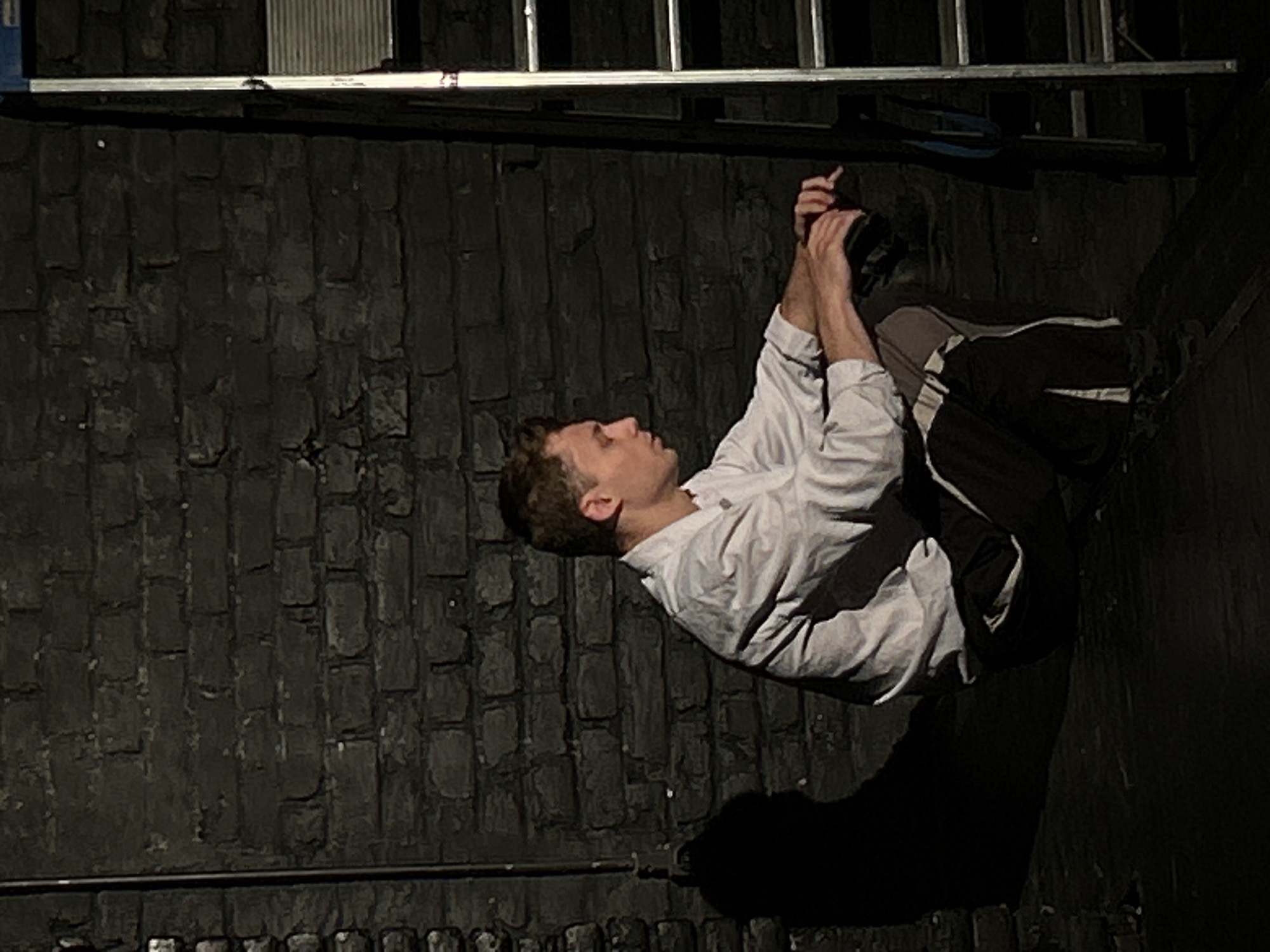 The image depicts a man hanging from a ladder, possibly indoors, with a dark, textured wall in the background. The man is wearing a light-colored long-sleeved shirt and dark pants with lighter-colored stripes on the sides. His hands are gripping the ladder. The ladder is made of metal. The location in the city of Unknown, Unknown, is unspecified.